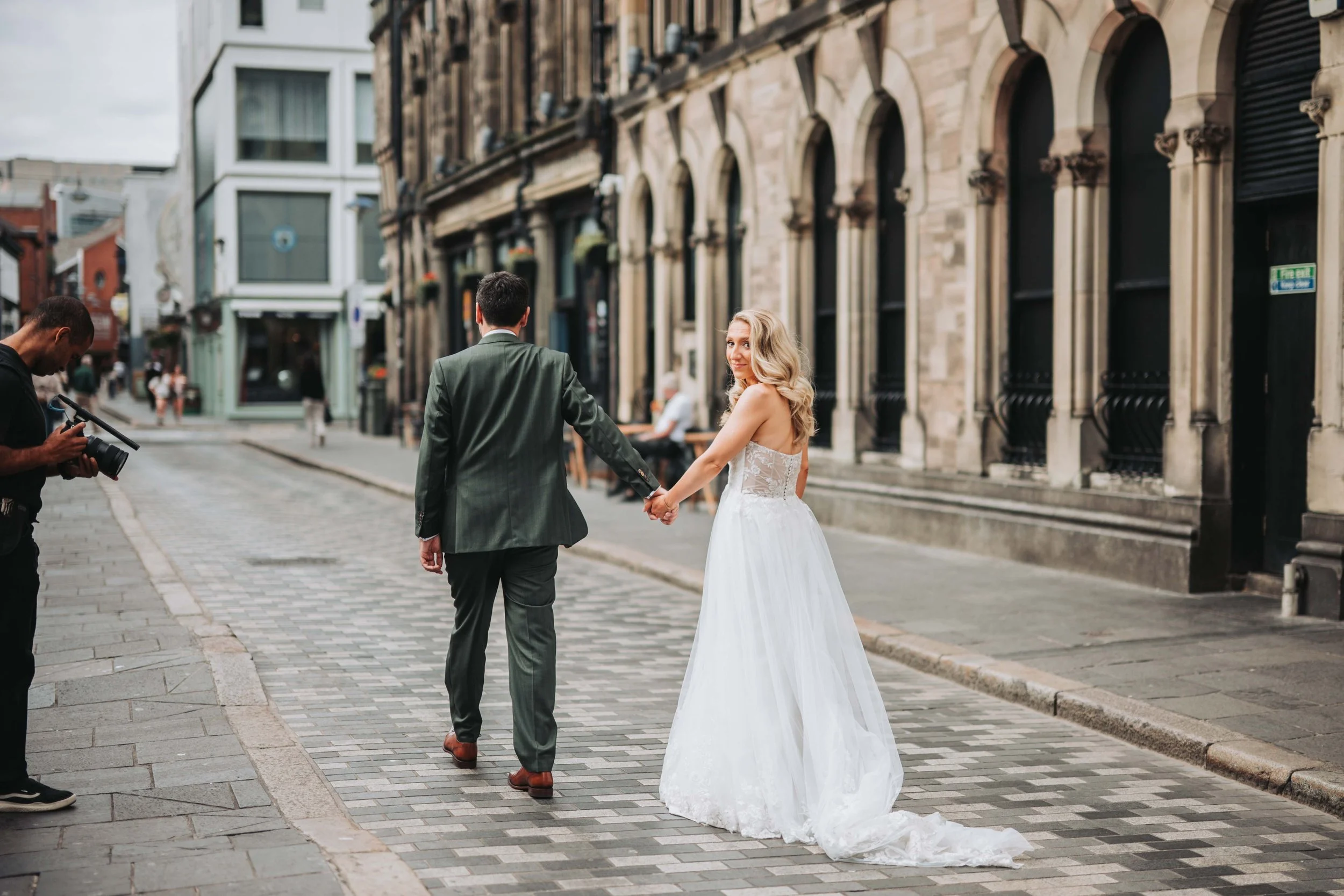 A couple, a man in a dark suit and a woman in a white wedding dress, holding hands and walking on a cobblestone street in an urban area with historic buildings. A photographer is standing nearby, taking pictures.