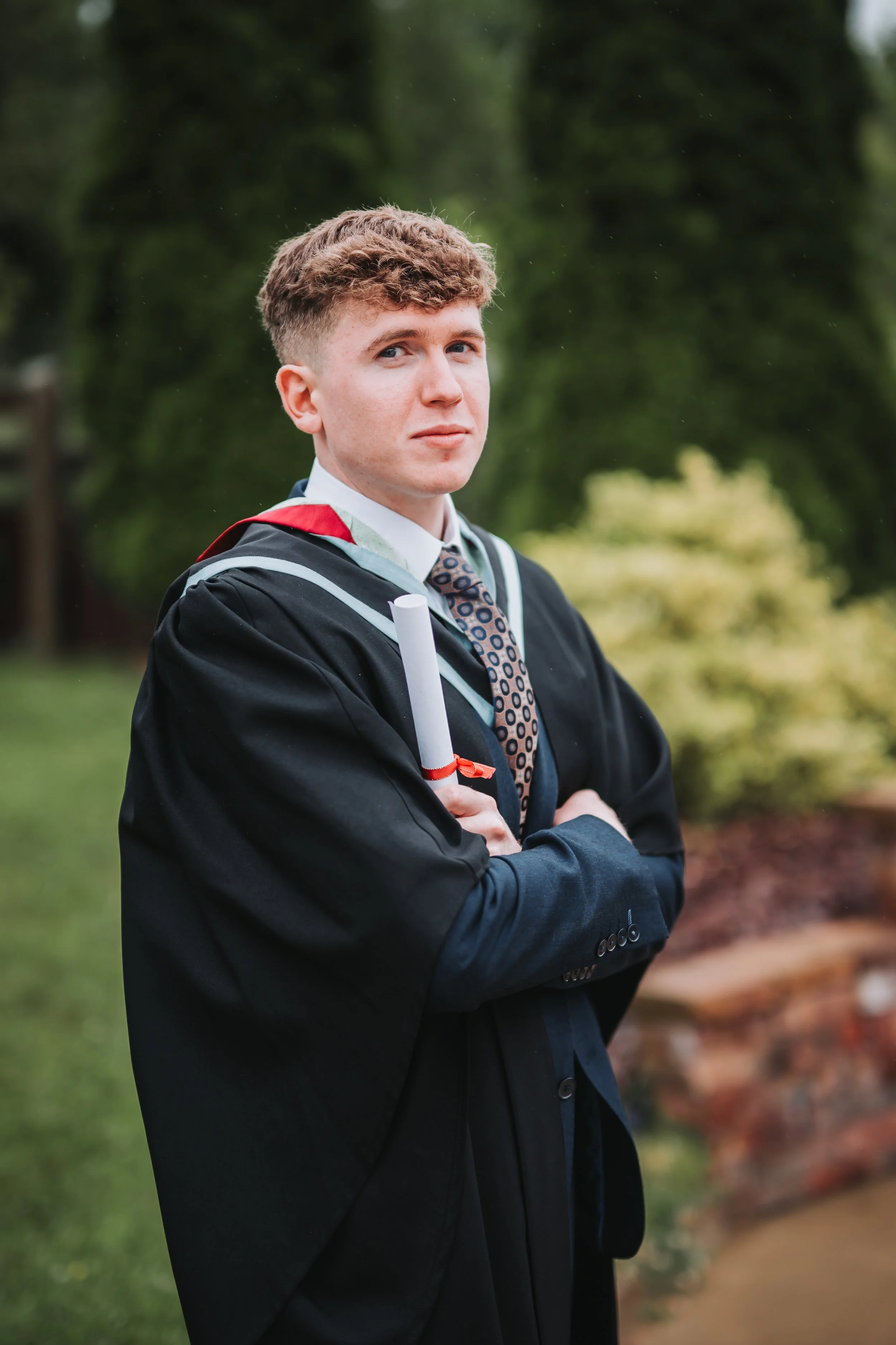 A young man in a graduation gown holding a diploma, standing outdoors with green trees and grass in the background.