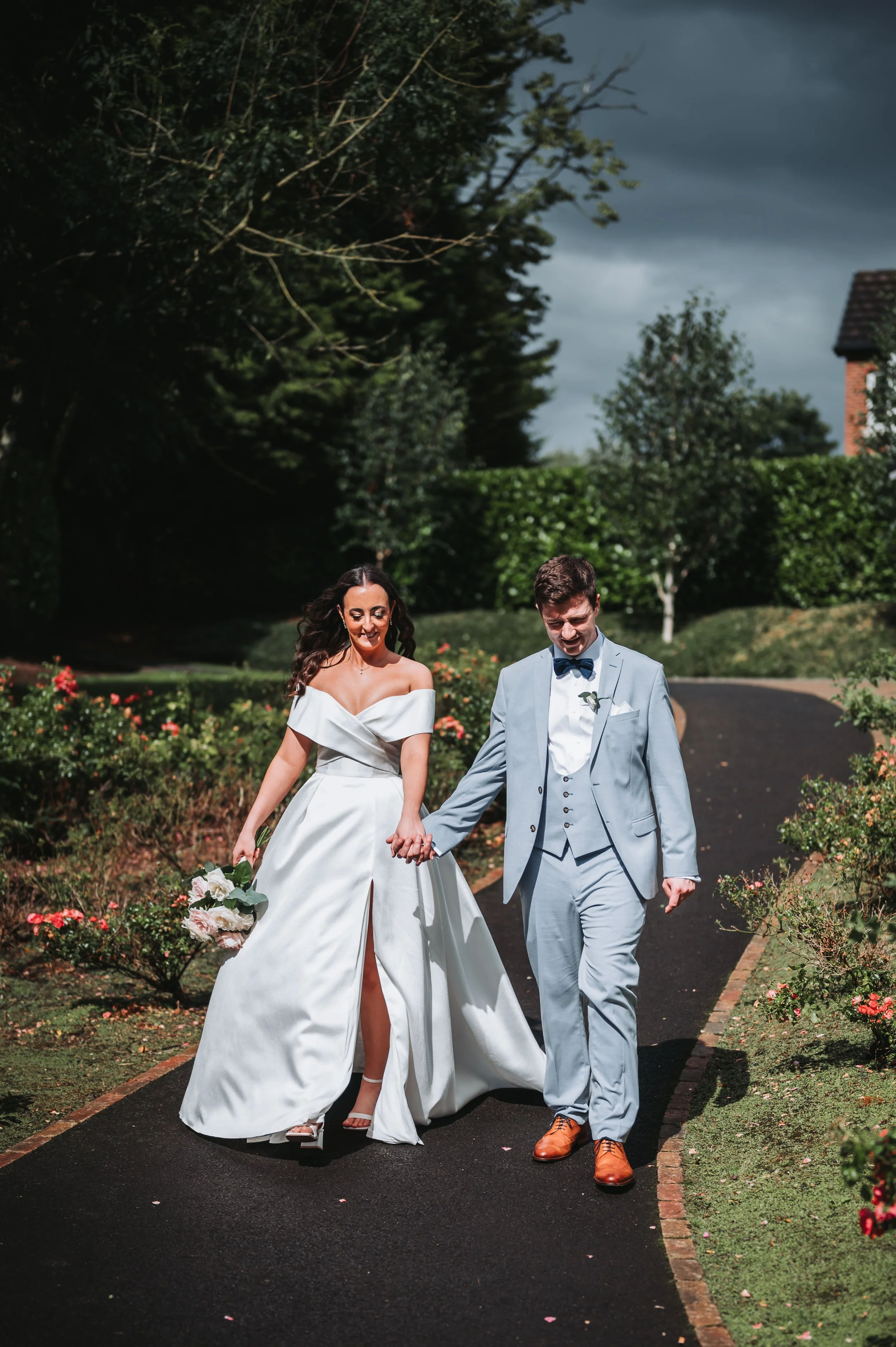 Bride and groom walking hand in hand on a garden path, smiling, with bridal gown and tuxedo, surrounded by greenery and flowers.