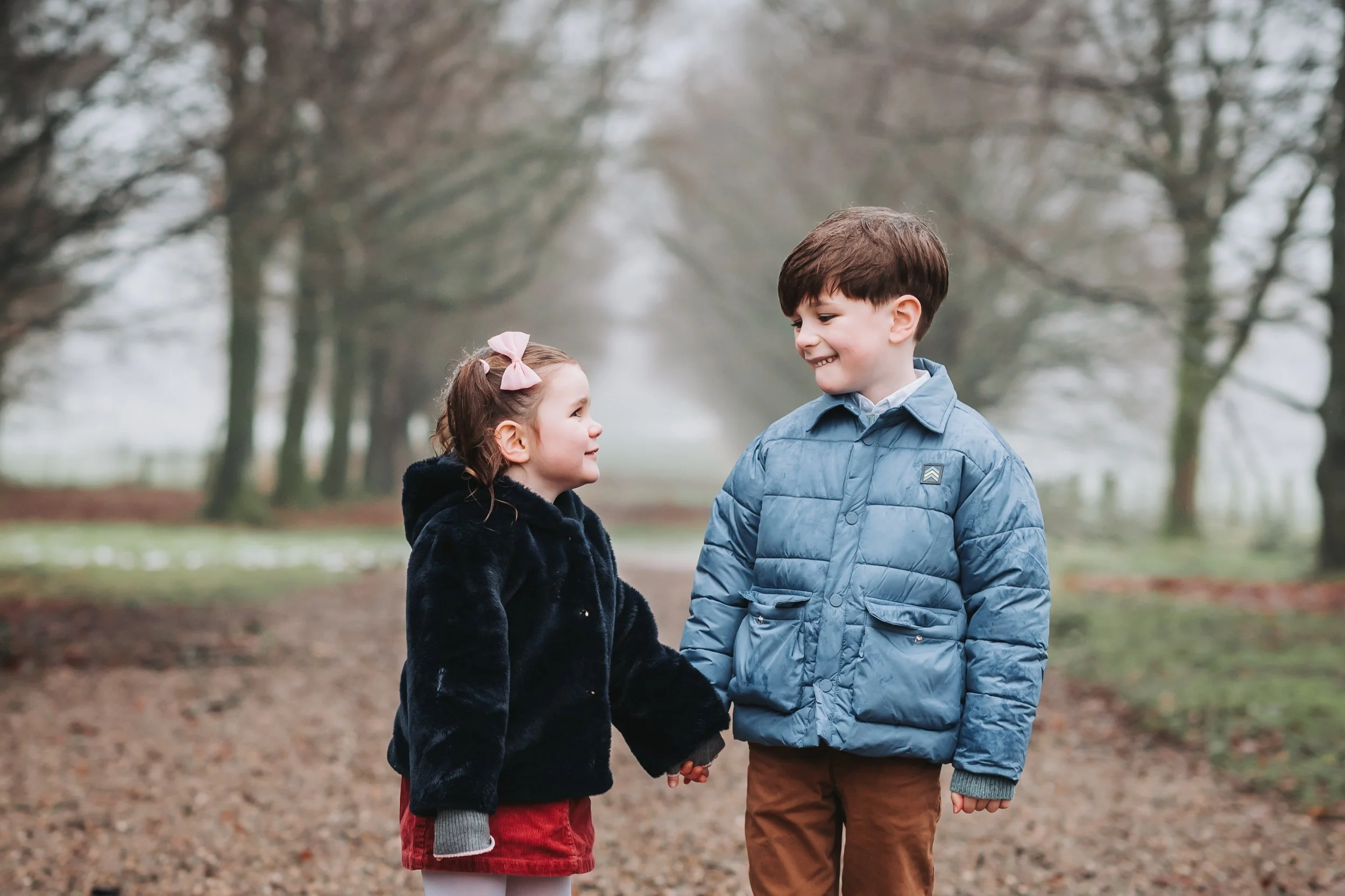 A young girl and boy holding hands and smiling at each other on a tree-lined outdoor path in a foggy setting.