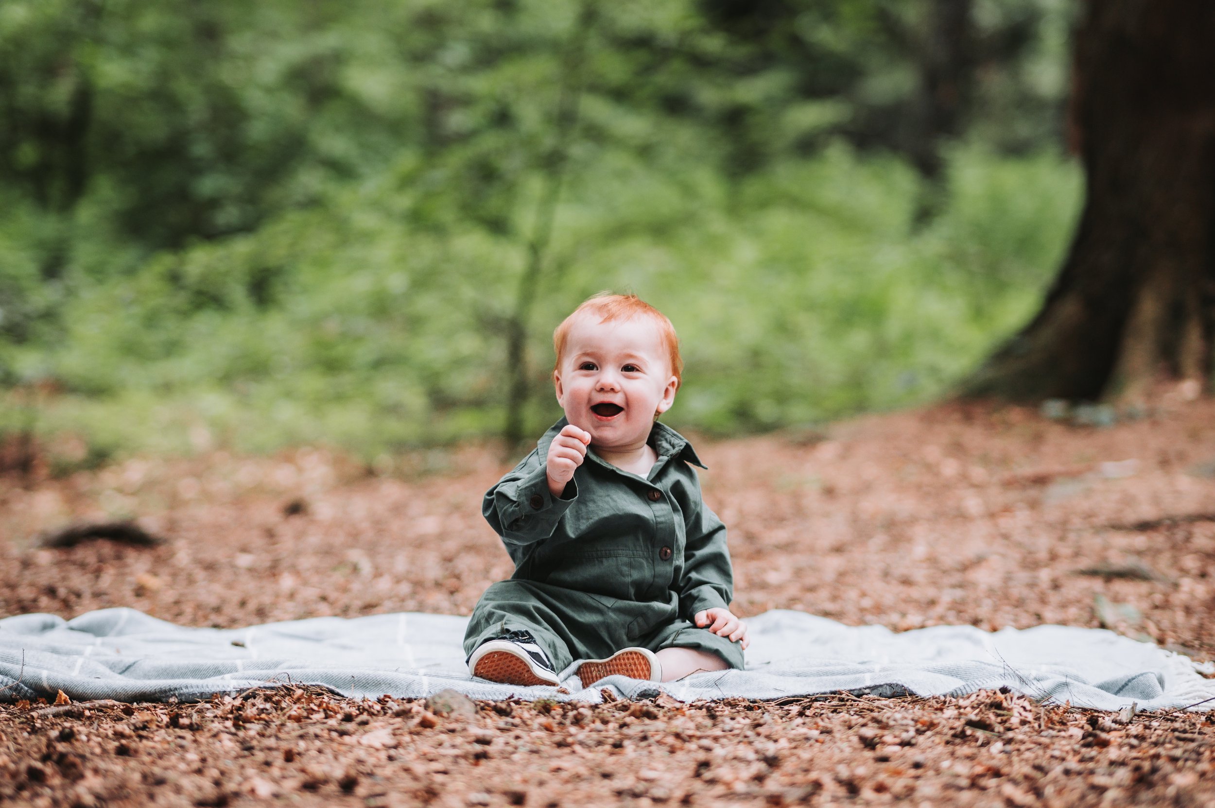 A happy baby with red hair sitting on a blanket in a wooded park