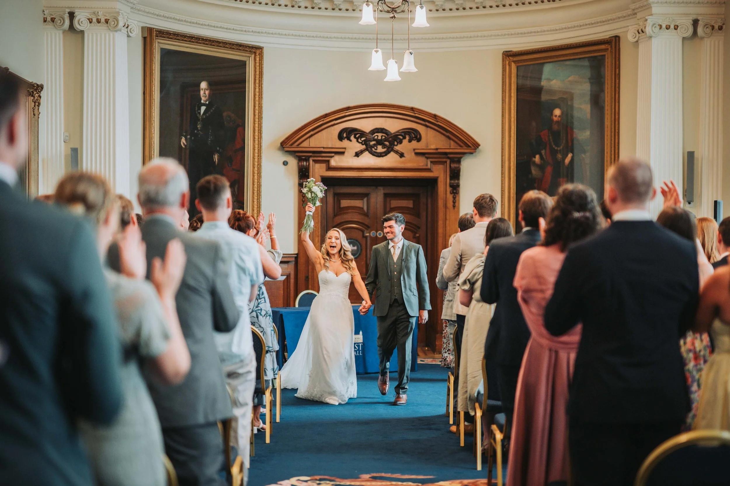 A bride and groom walking down the aisle of a church, holding hands, with the bride raising a bouquet in her right hand. They are smiling and surrounded by guests who are applauding and cheering.