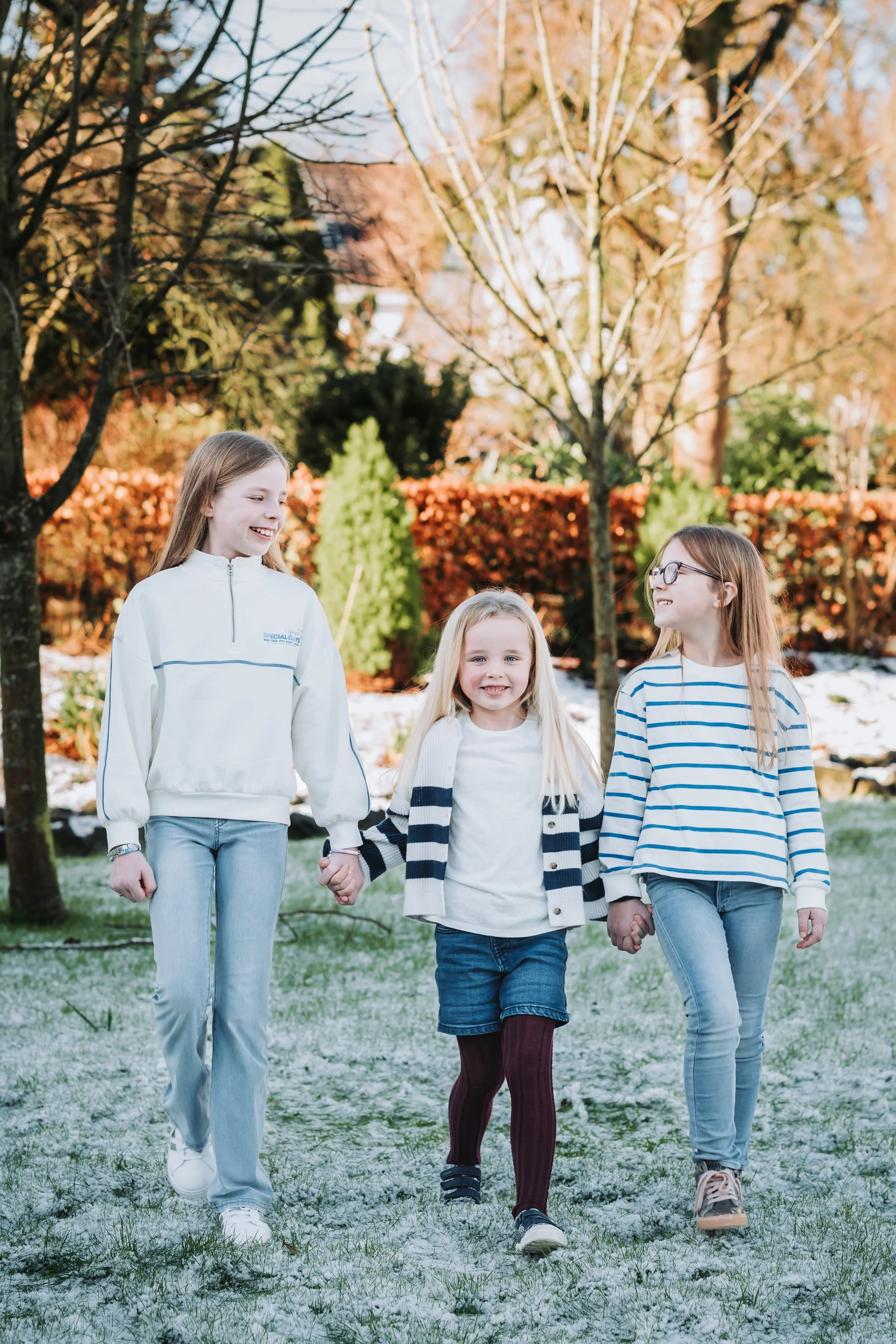 Three young girls walking outside on a cold day, holding hands and smiling, with trees and autumn foliage in the background.
