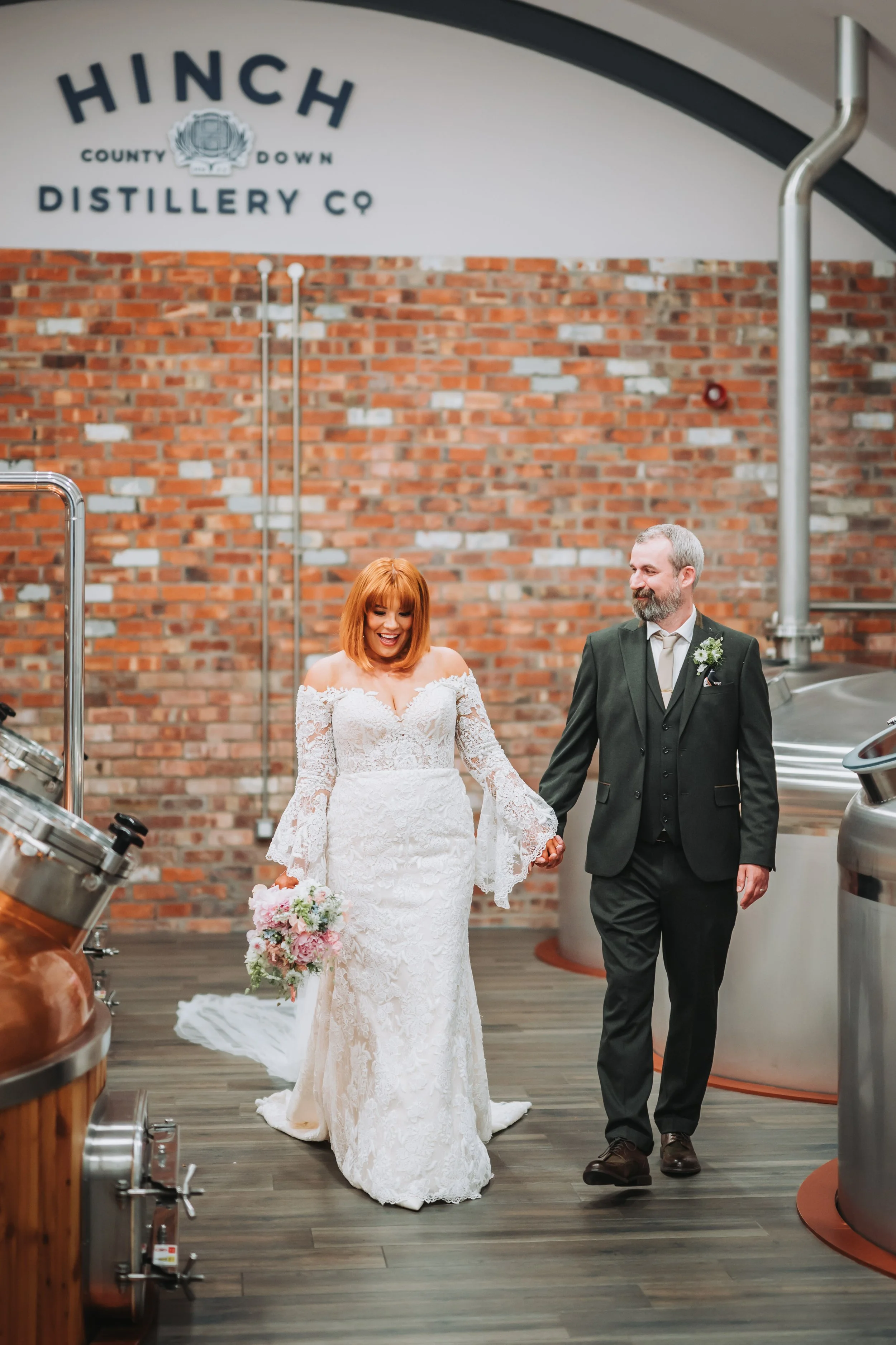 A bride in a white lace wedding dress holding a bouquet, walking hand-in-hand with a groom in a dark suit inside a distillery with a brick wall and distillation equipment.