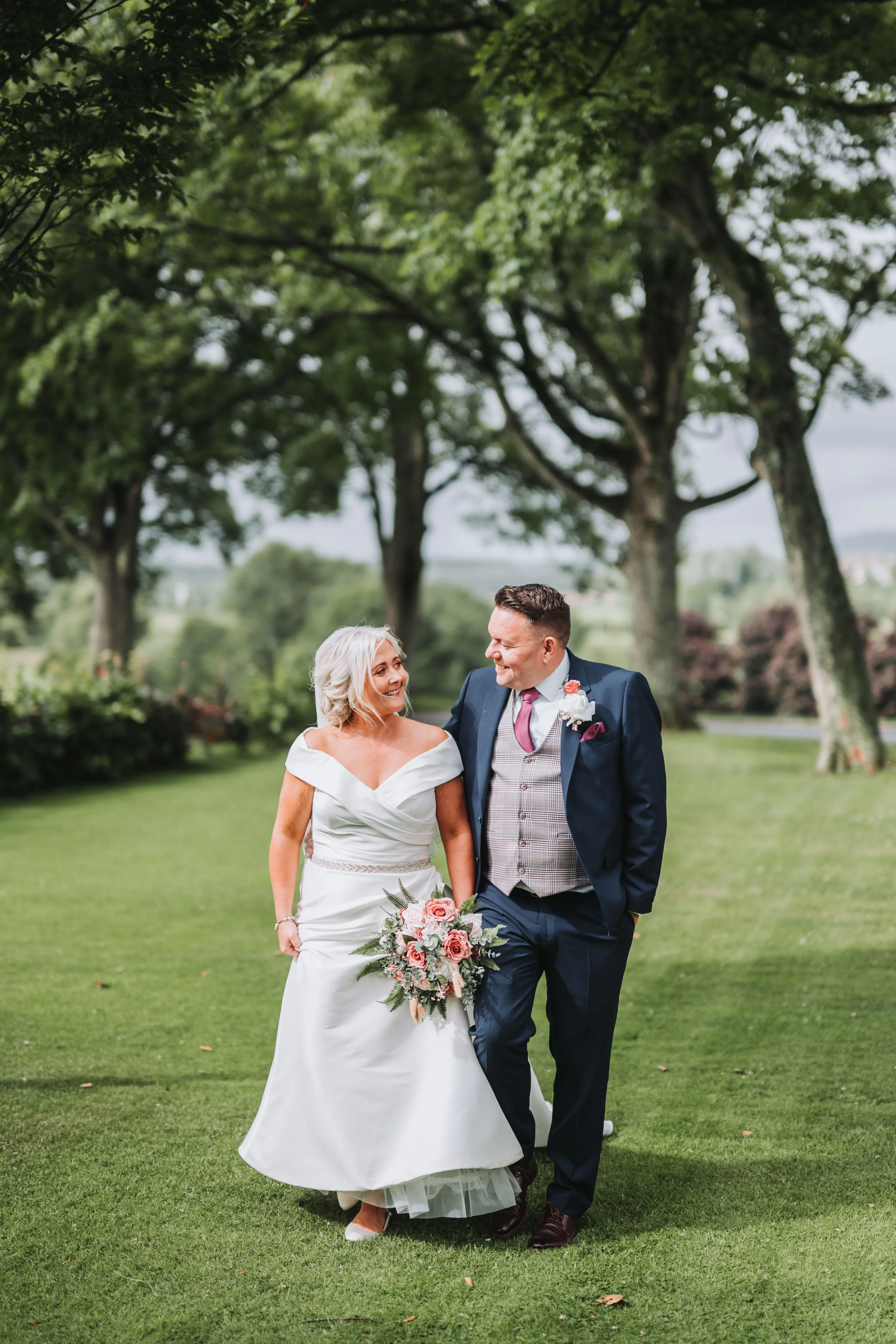 A bride and groom walking and smiling on a grassy lawn with trees in the background during their wedding celebration.