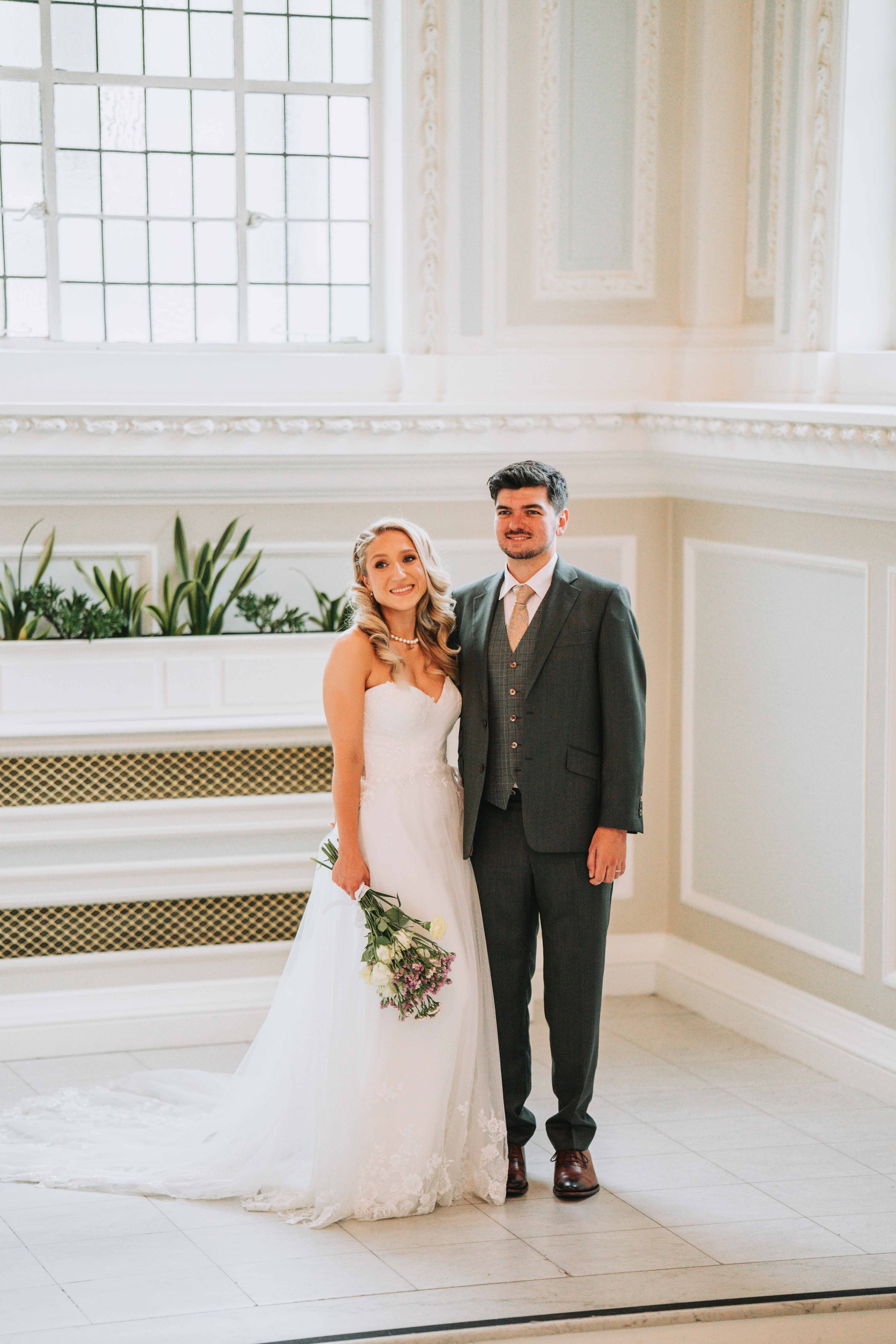 A bride and groom standing inside a bright, elegant room with large windows and decorative molding. The bride is holding a bouquet, and they are smiling at the camera.