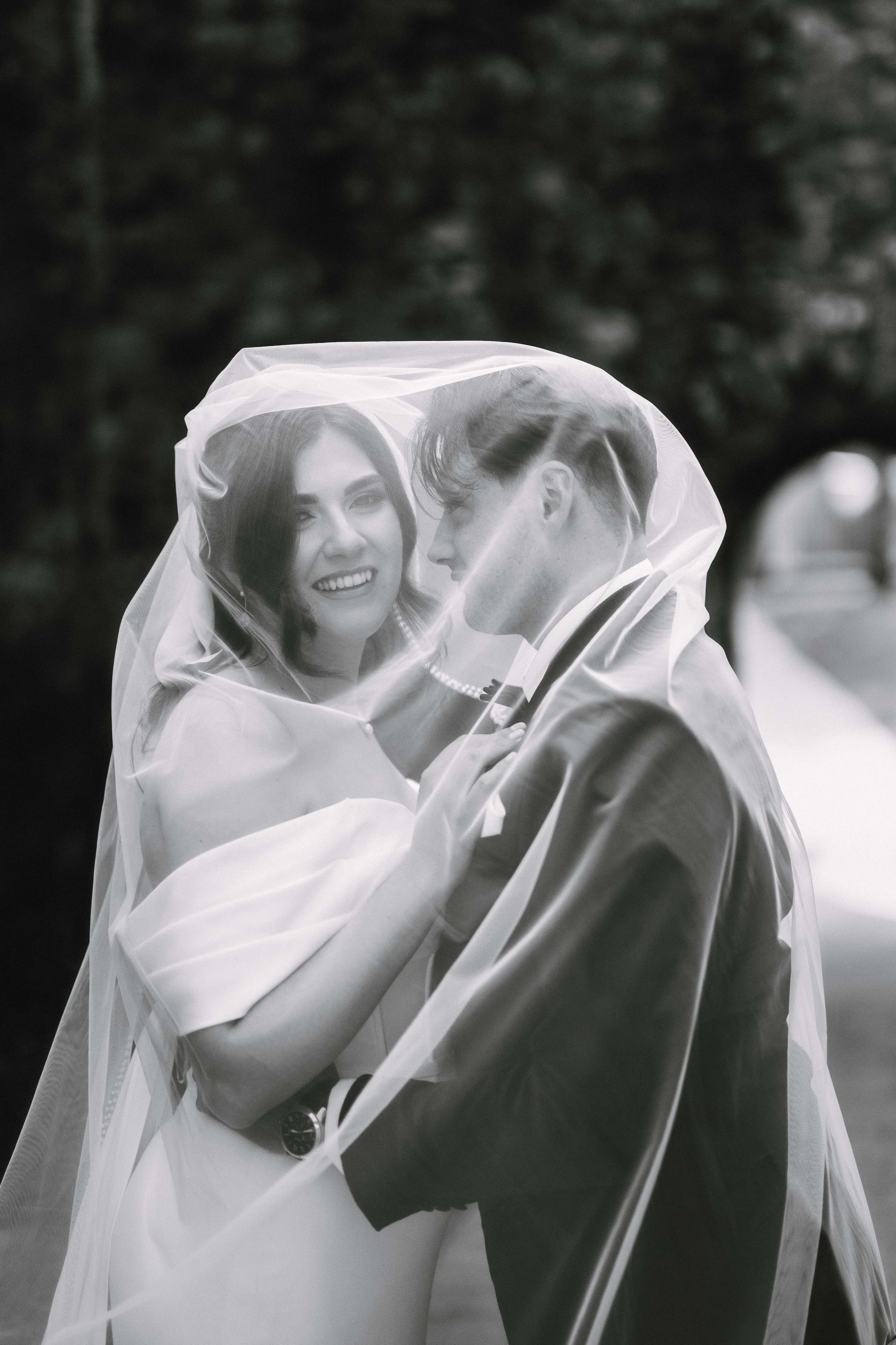 A bride and groom under a sheer veil, smiling and sharing a close moment outdoors.