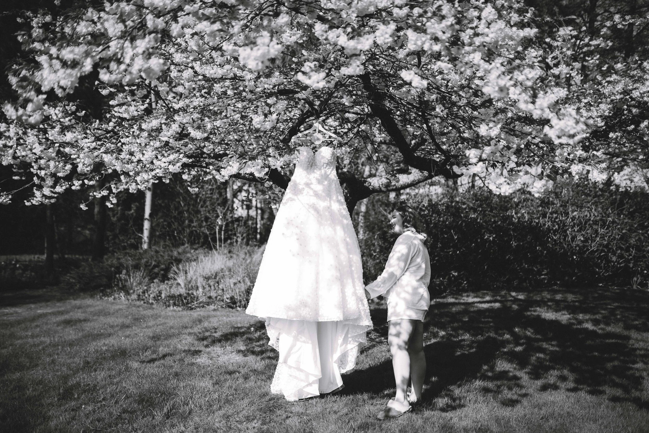 A woman holding hands with a person on stilts dressed in a wedding gown under a blooming tree, black and white photograph.
