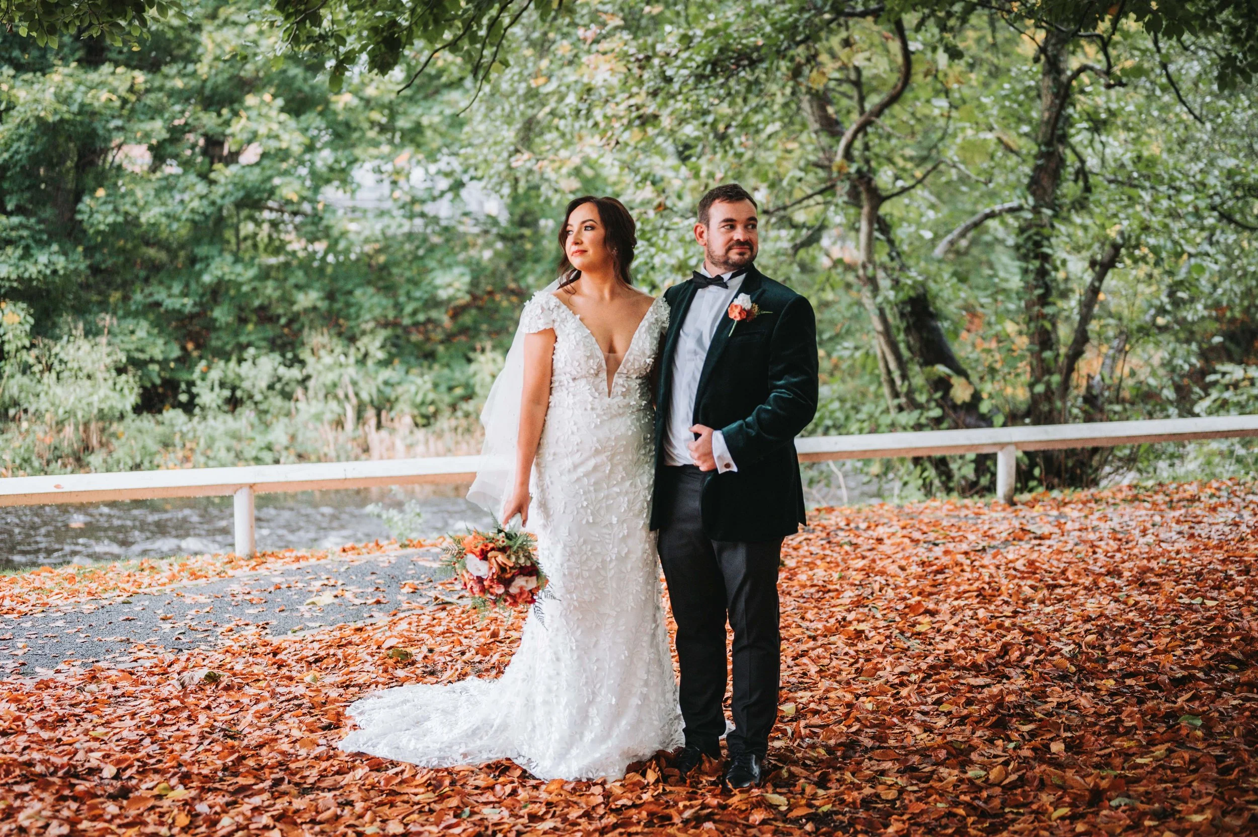 Bride and groom standing outdoors on a path covered with fallen orange and brown leaves, surrounded by green trees, during a wedding.