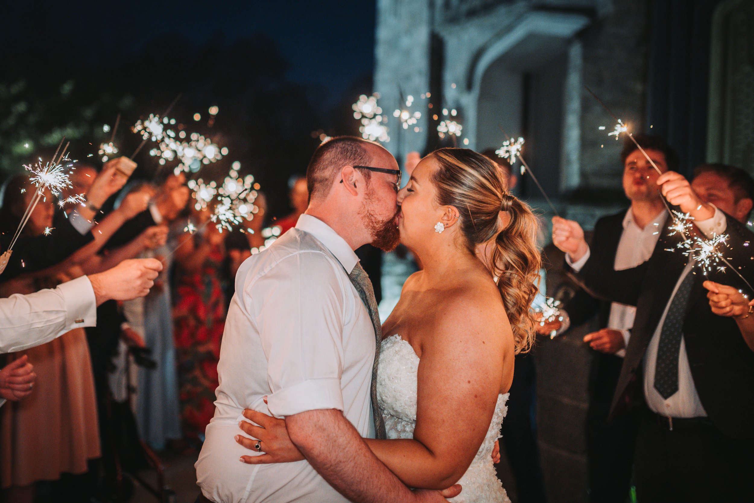 A newlywed couple sharing a kiss at their wedding reception surrounded by guests holding sparklers.