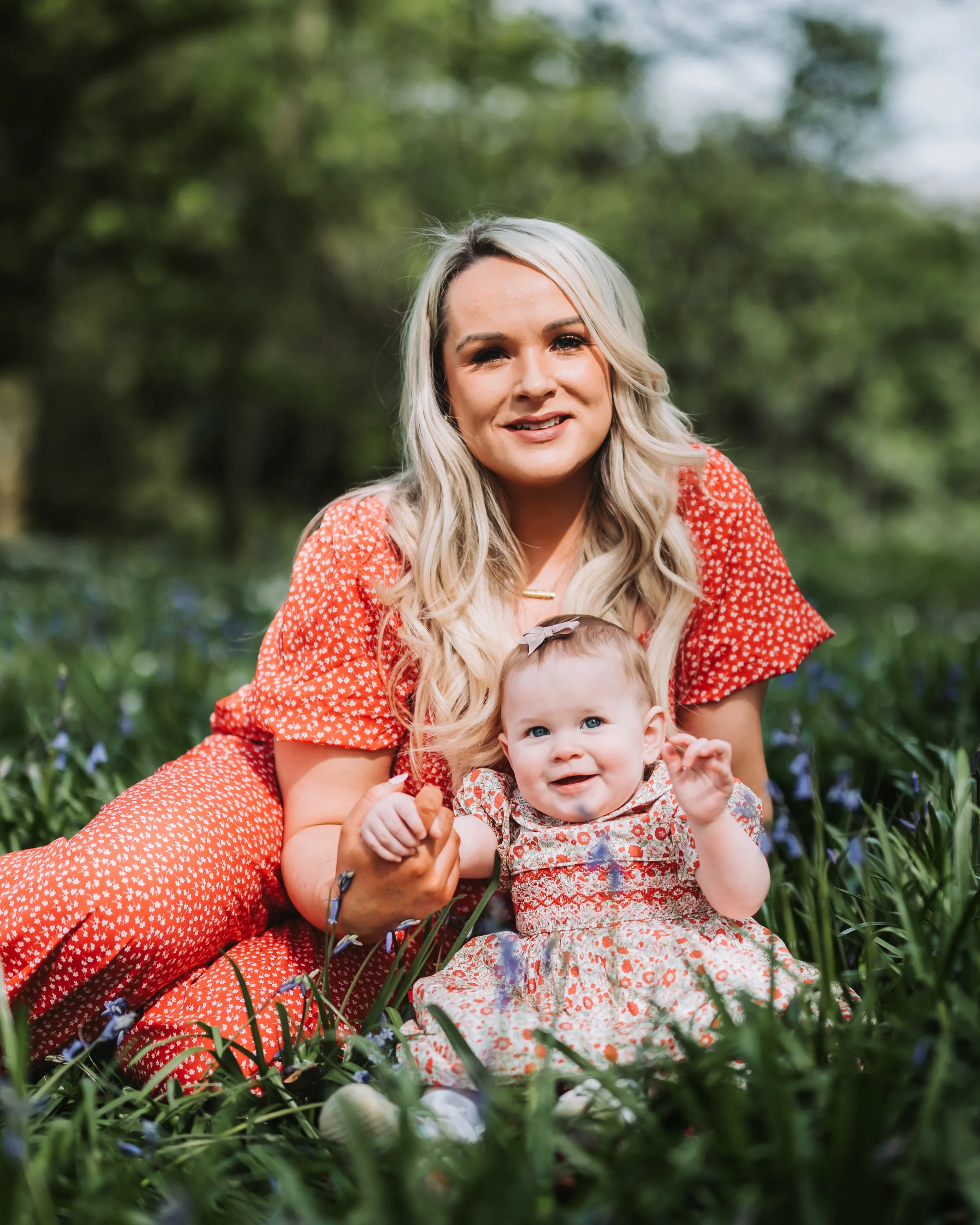 A woman and a young girl sitting outdoors on the grass, surrounded by greenery and small flowers, both smiling at the camera.