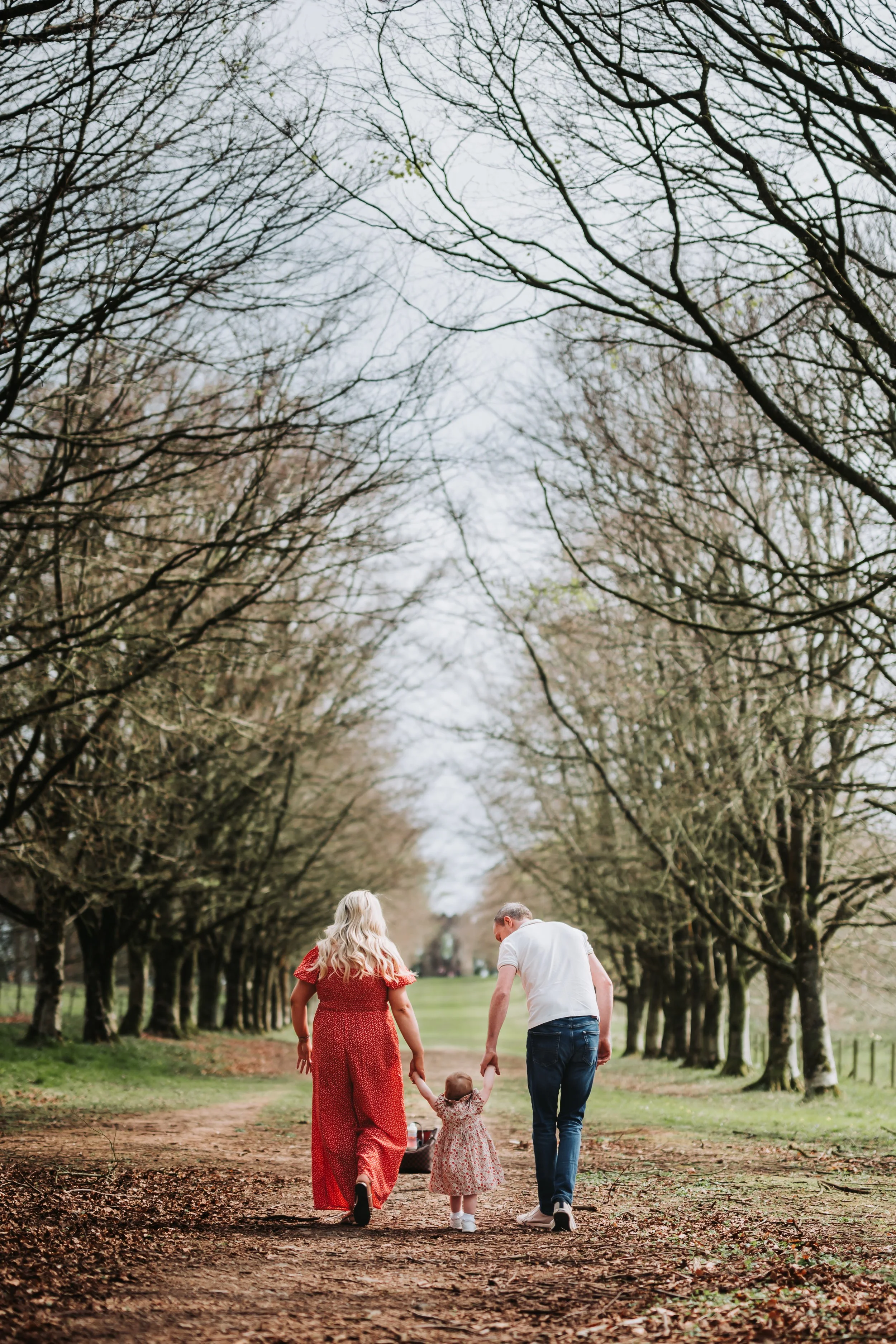 A family of three walking down a dirt path lined with bare trees, holding a child's hands, during daytime.