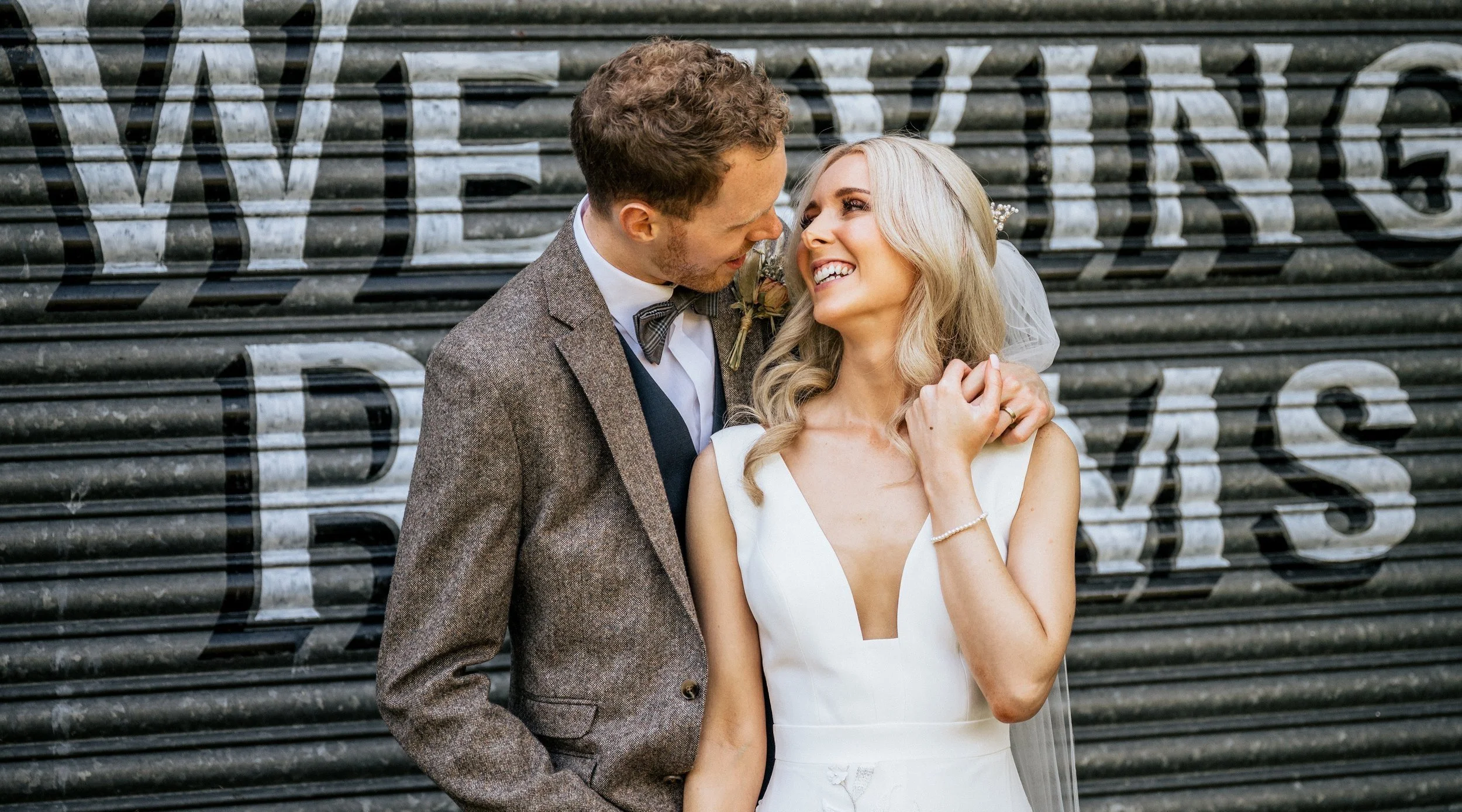 A bride in a white dress and a groom in a brown suit smiling at each other in front of a metal shutter with large text.