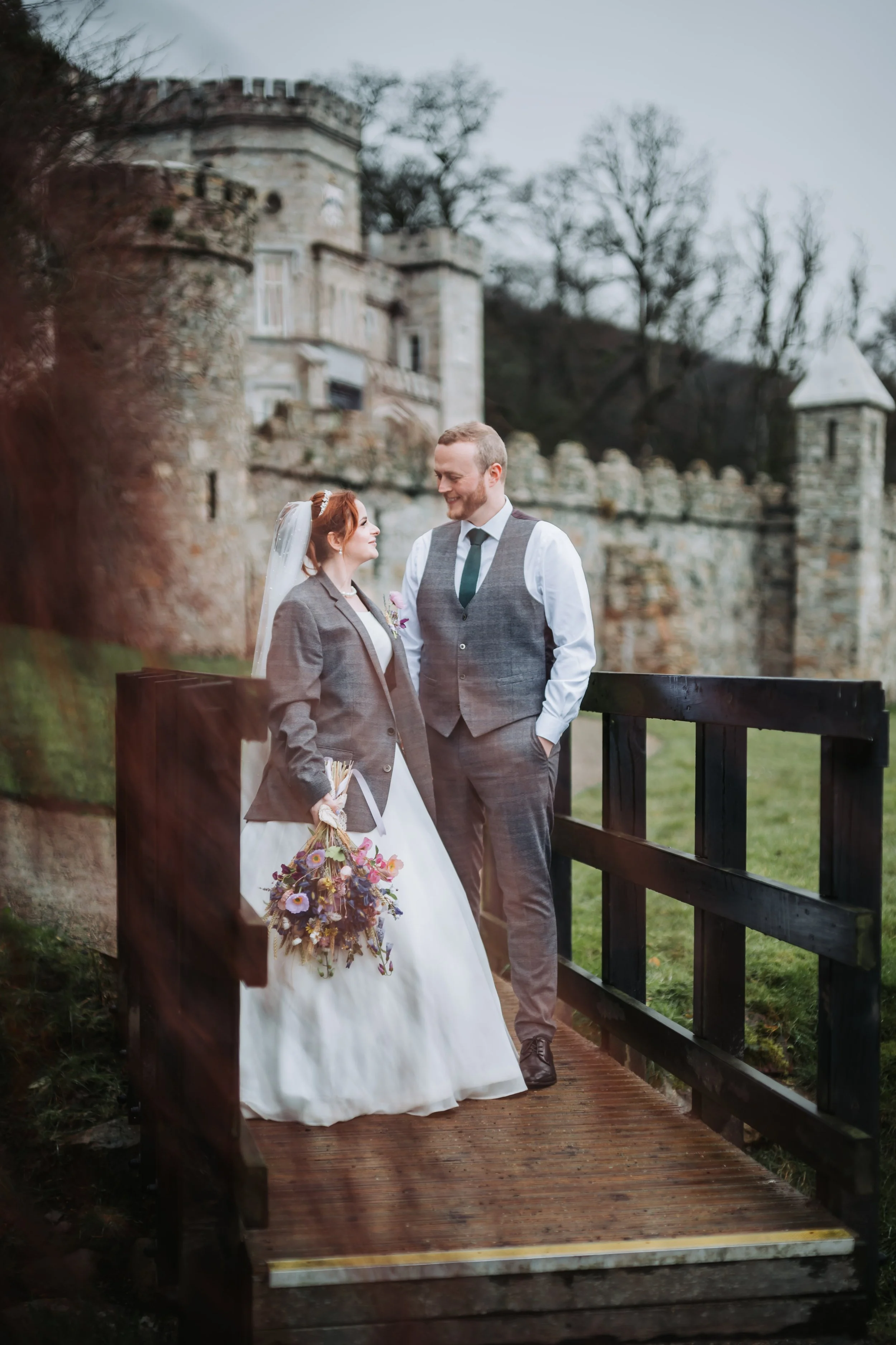A bride and groom standing on a small wooden bridge outdoors, smiling at each other with a castle in the background.