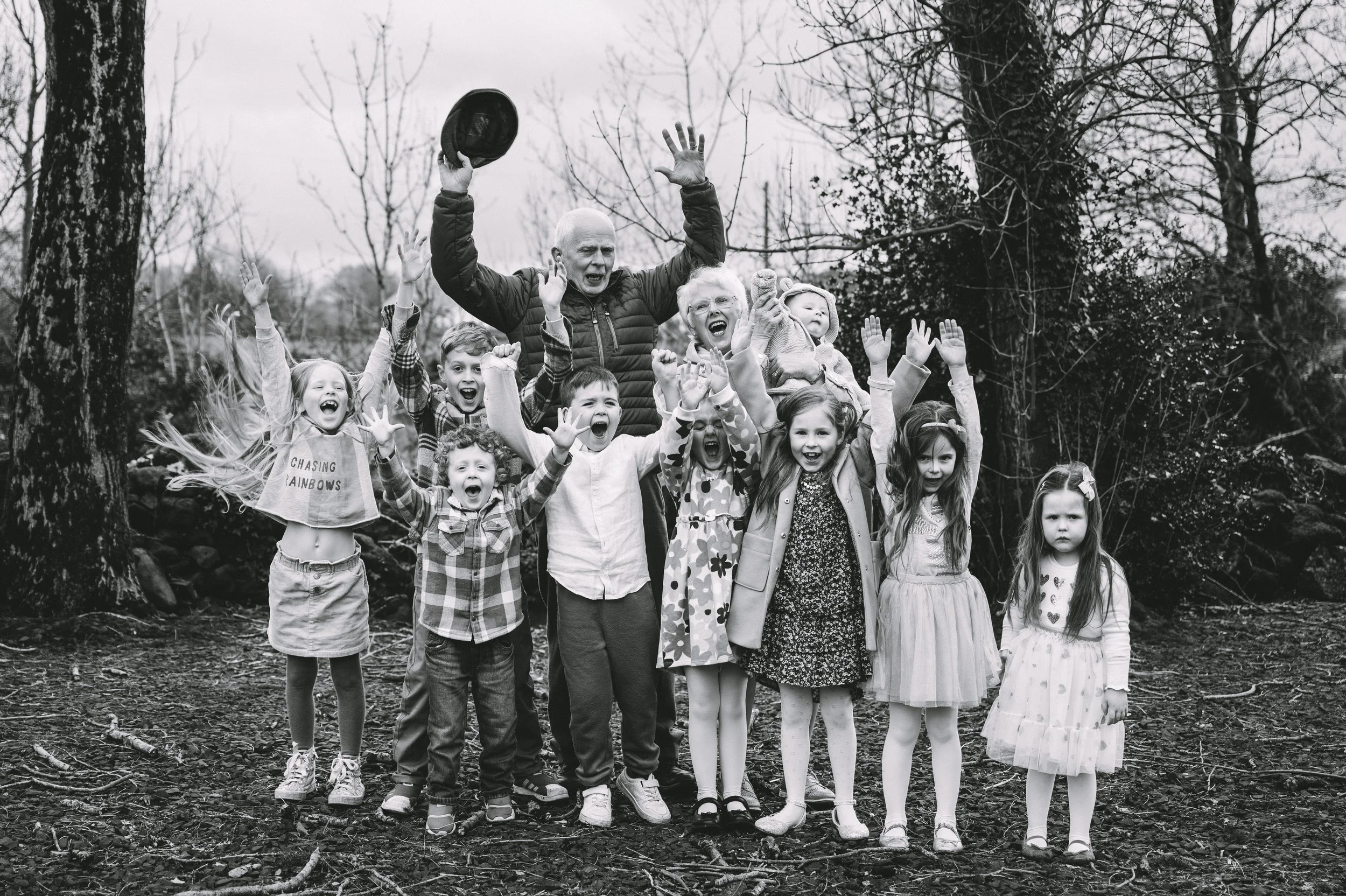 Black and white photo of a group of children and an adult outdoors, all cheerfully raising their hands, with trees in the background.