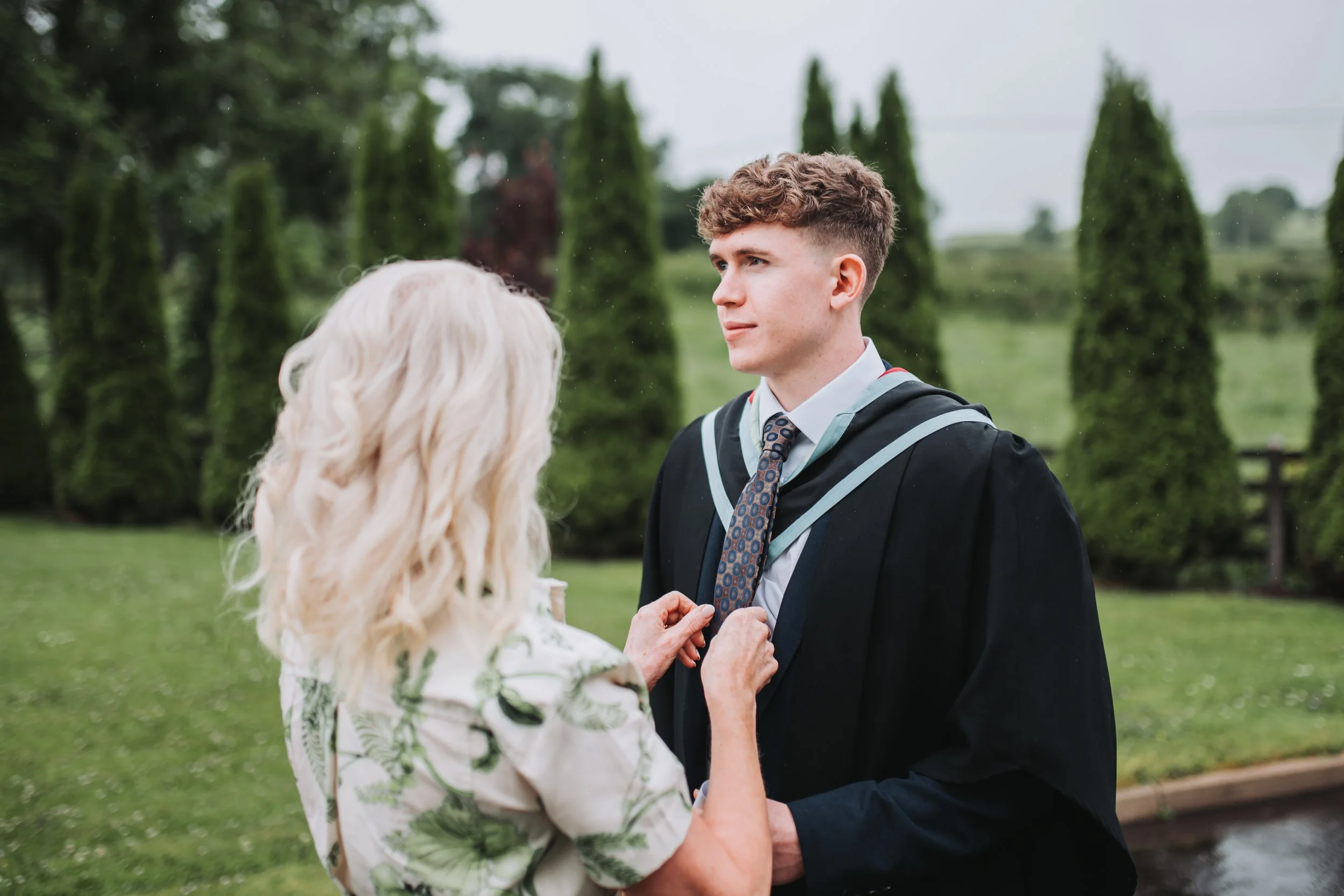 A young man in a graduation gown and tie being awarded a diploma by a woman with blonde curly hair outdoors, with trees and a cloudy sky in the background.