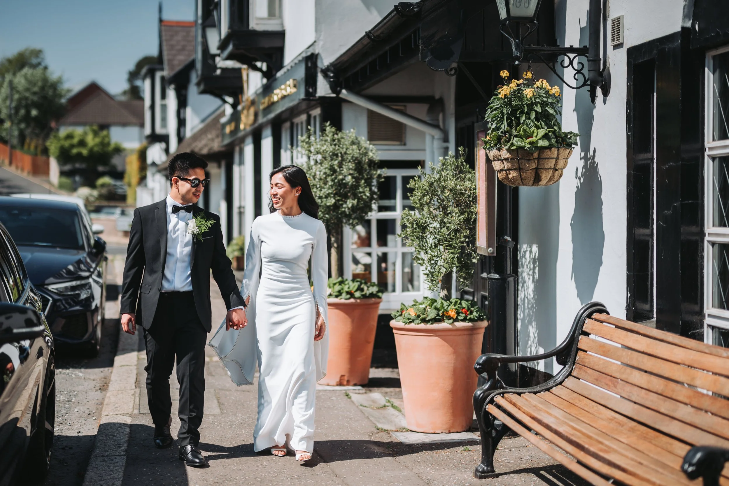 A bride and groom walking hand in hand along a sidewalk on a sunny day, smiling and looking at each other. The groom is wearing a black tuxedo and sunglasses, while the bride is in a long white wedding dress. There are potted plants and a building wi