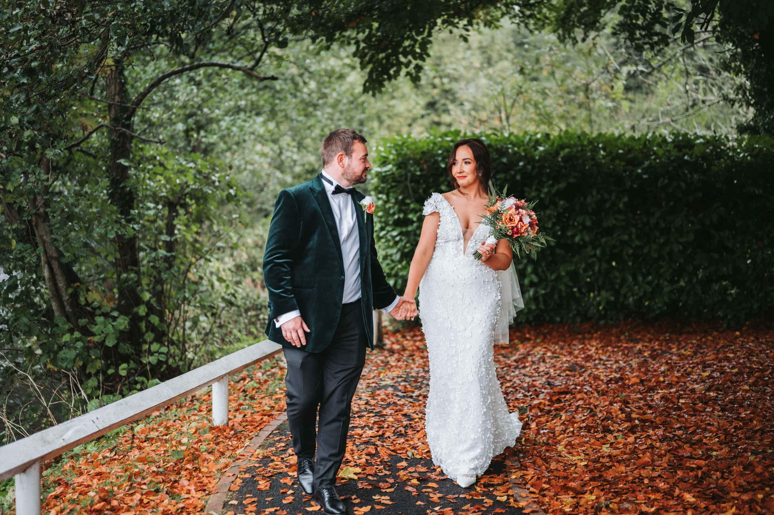 A bride and groom holding hands and walking through a leaf-covered path in a park on their wedding day. The bride is wearing a white lace wedding dress and holding a bouquet, while the groom is dressed in a dark green velvet blazer and black trousers