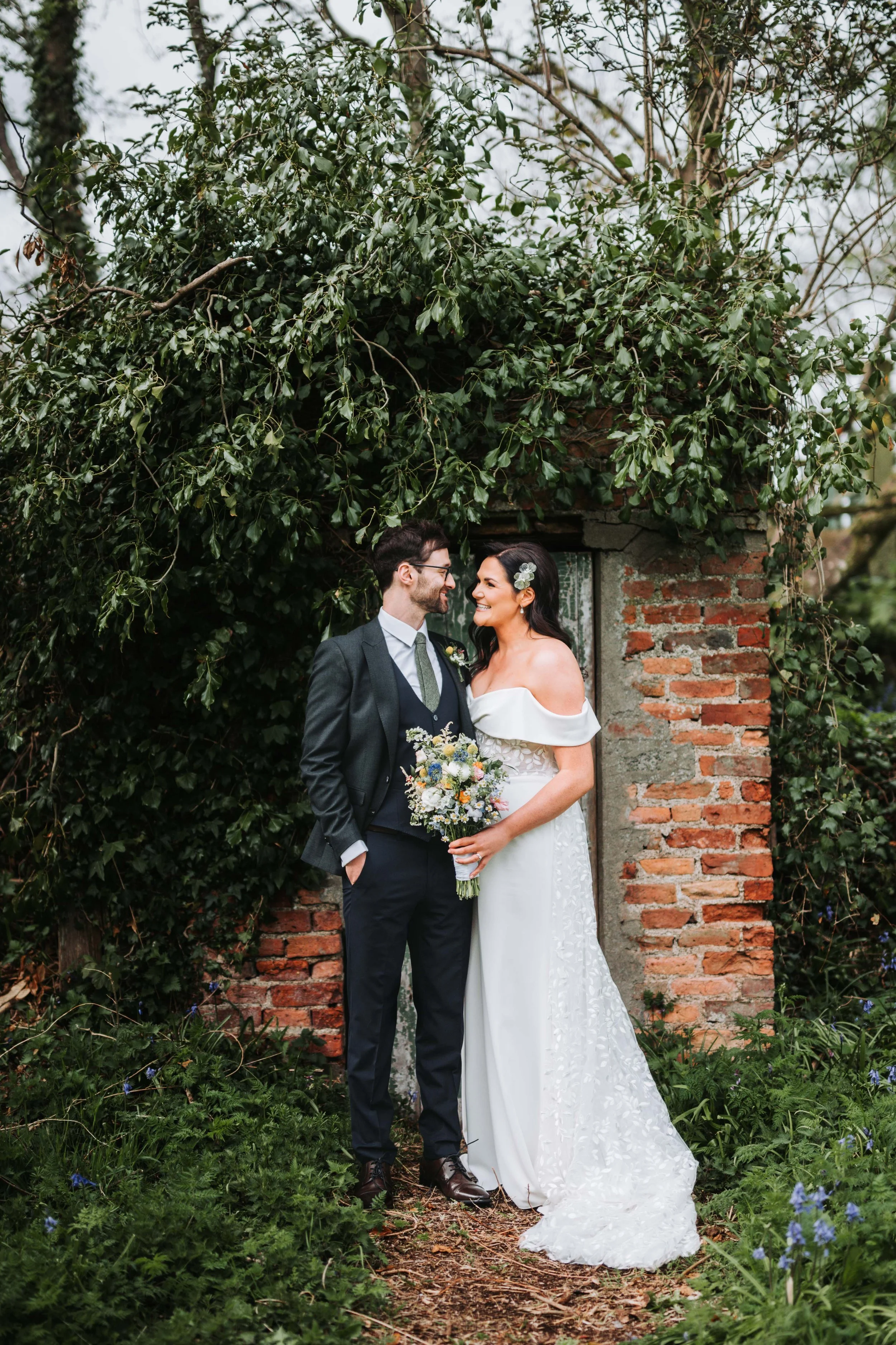 A bride and groom standing close together outdoors by a brick and stone structure, smiling at each other. The bride wears a white off-shoulder wedding dress with floral embroidery, and holds a bouquet of flowers. The groom wears a dark suit, vest, an