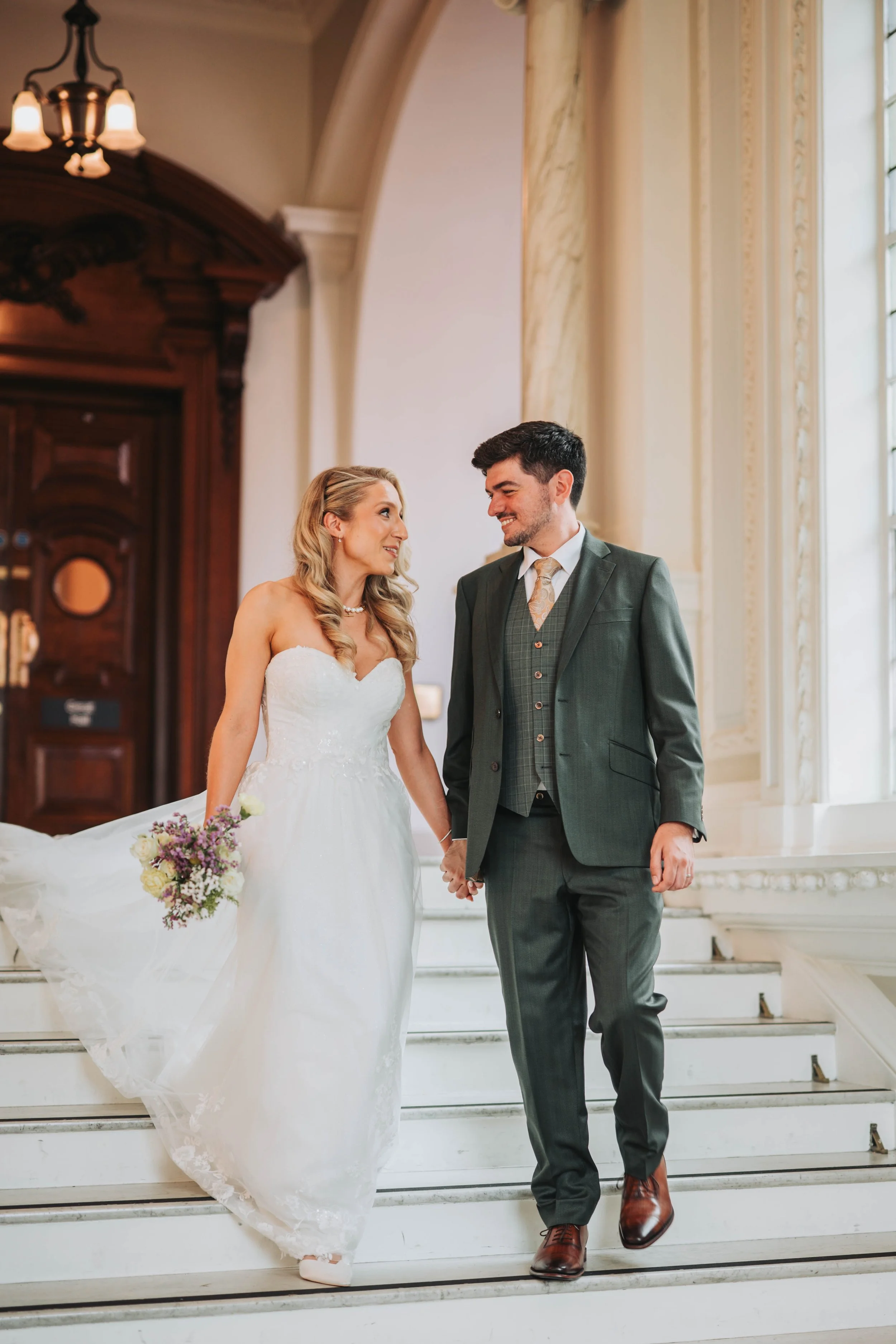 Bride and groom holding hands on a staircase during their wedding in a grand, historic building.