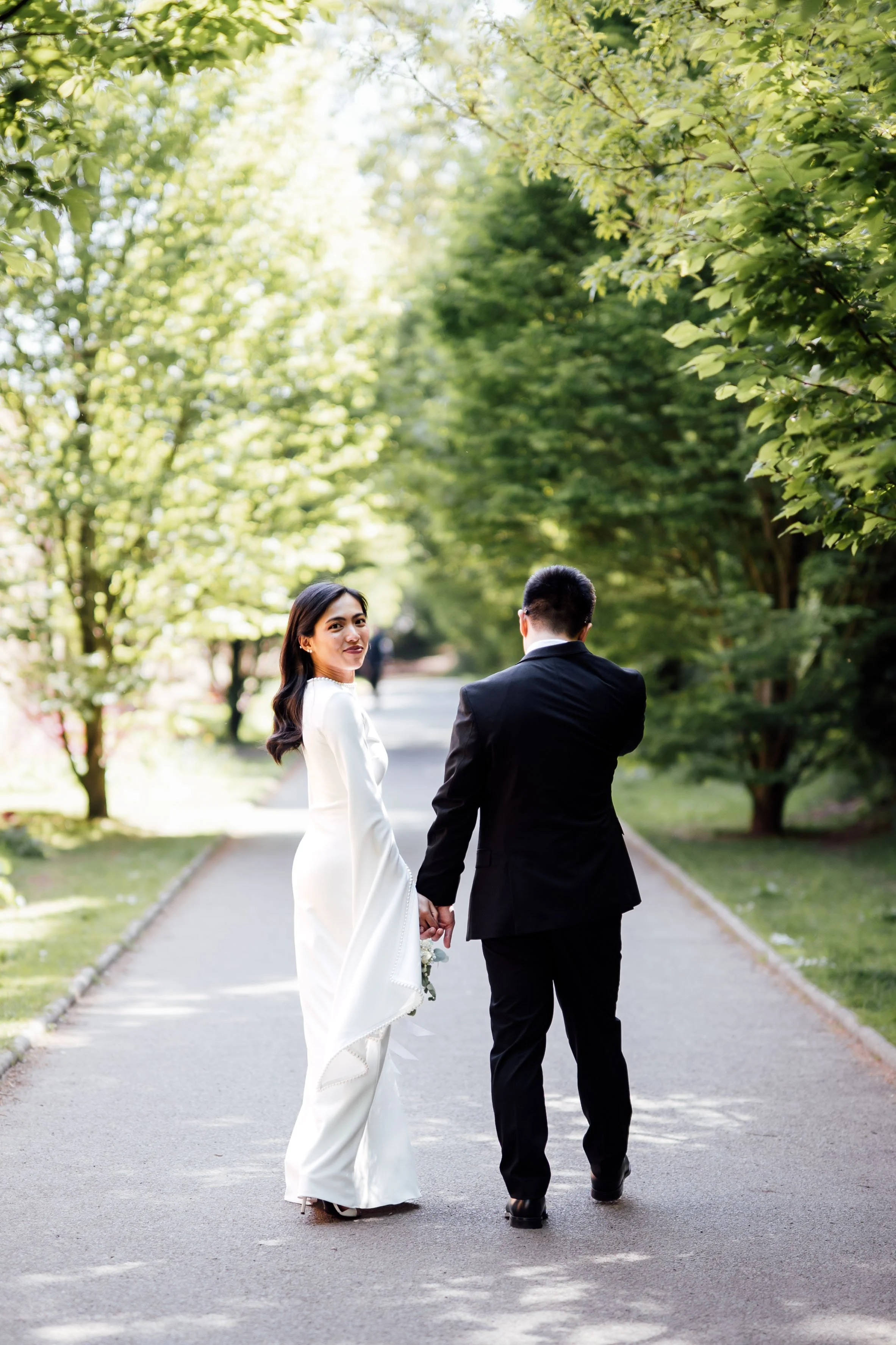 A couple in wedding attire holding hands and walking along a tree-lined pathway in a park or garden during daytime.