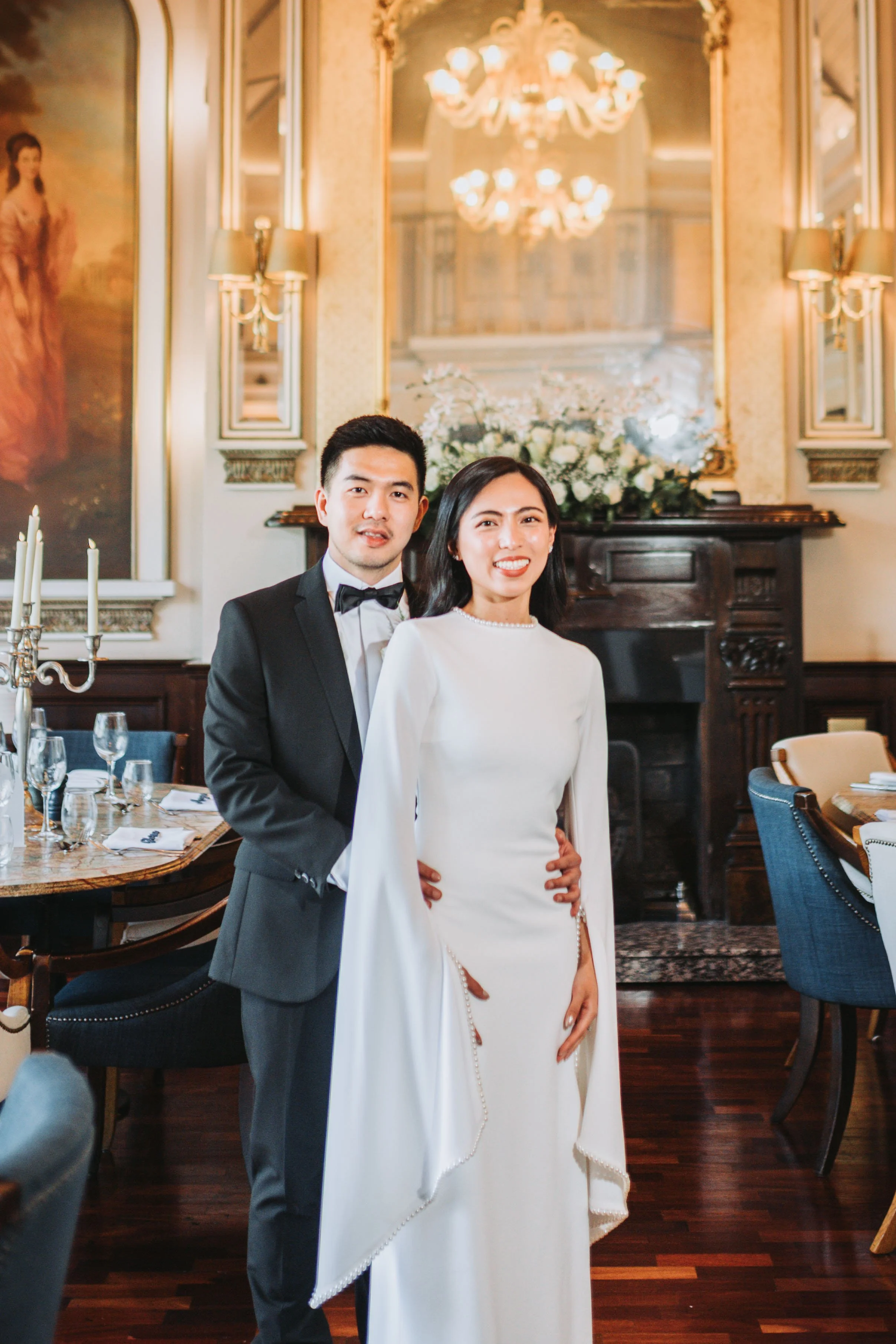A newlywed couple standing inside an elegant, richly decorated room with a fireplace, large mirror, and floral arrangements. The groom is wearing a black tuxedo with a bow tie, and the bride is dressed in a white gown with long sleeves and pearl embe