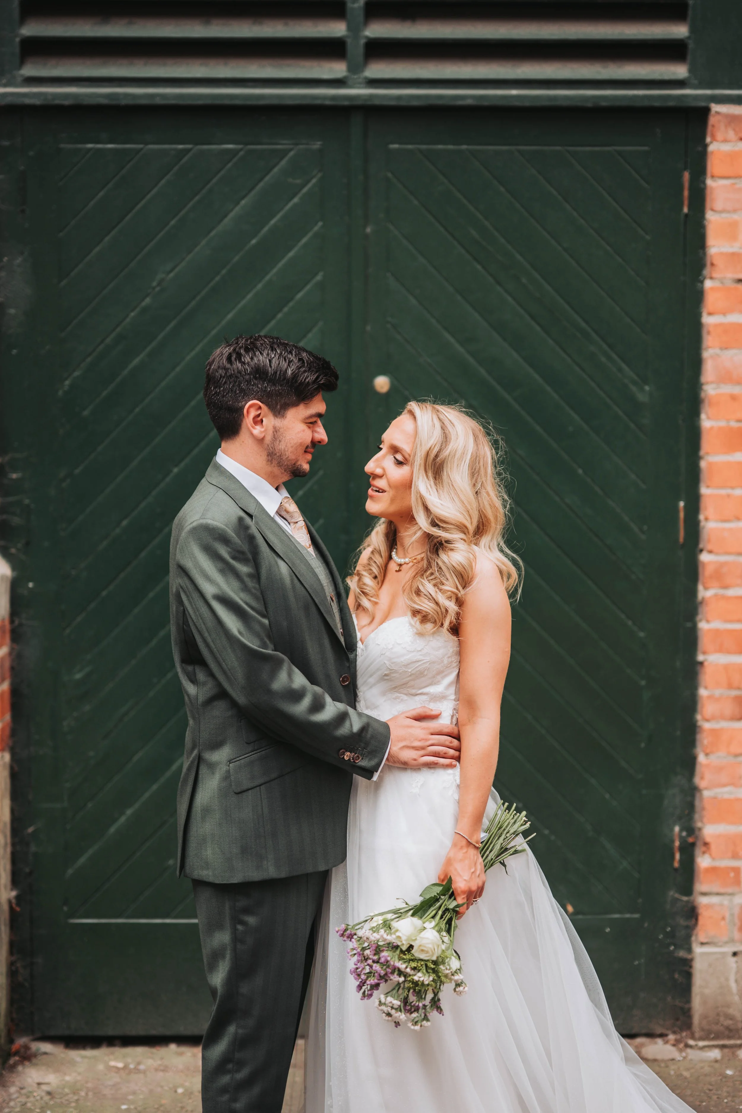 A bride and groom standing close together outside, smiling at each other. The bride is holding a bouquet of white and purple flowers. They are near a large green gate with brick walls on each side.