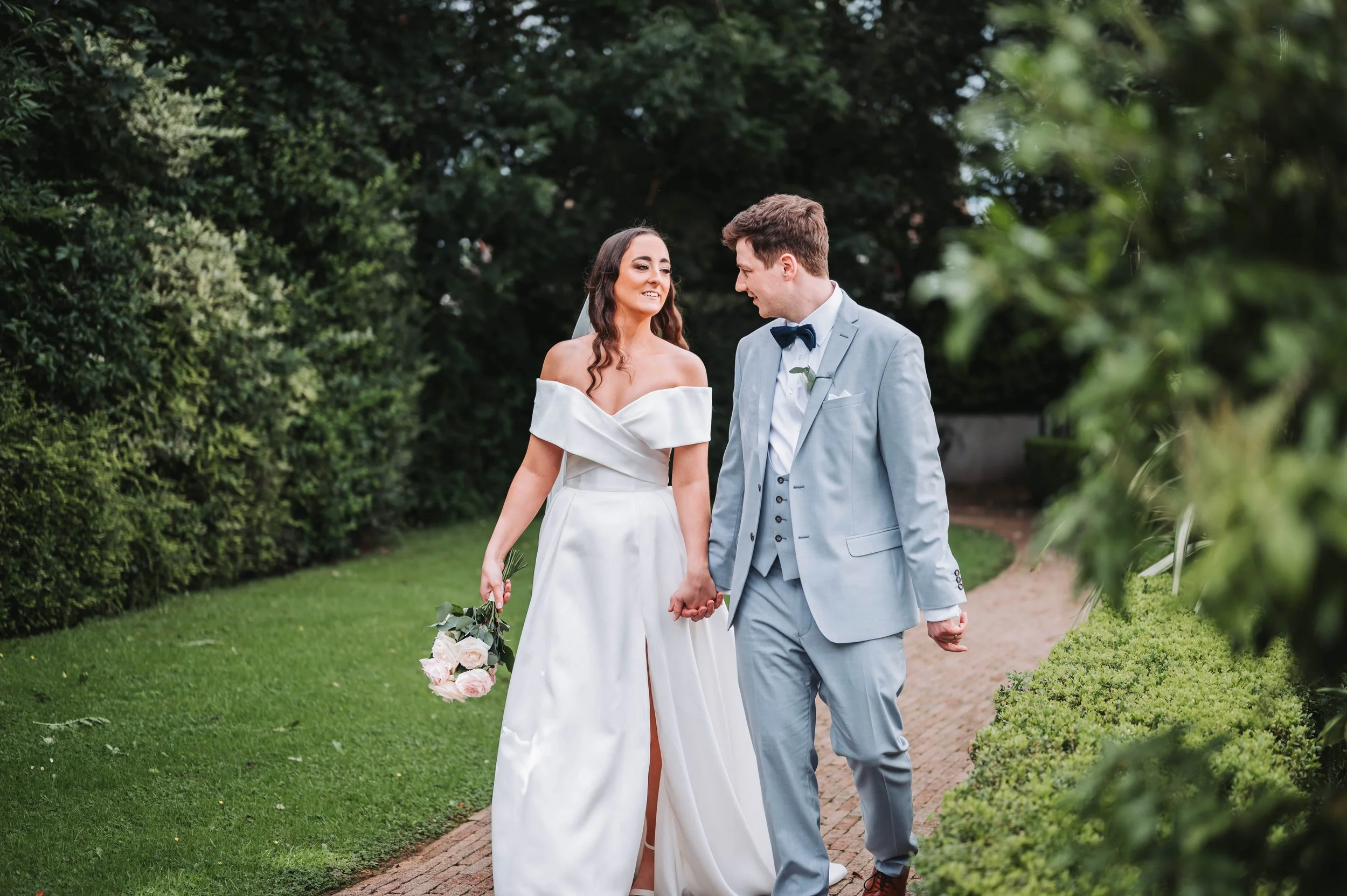 A bride and groom holding hands and walking together outdoors, surrounded by greenery, during their wedding.