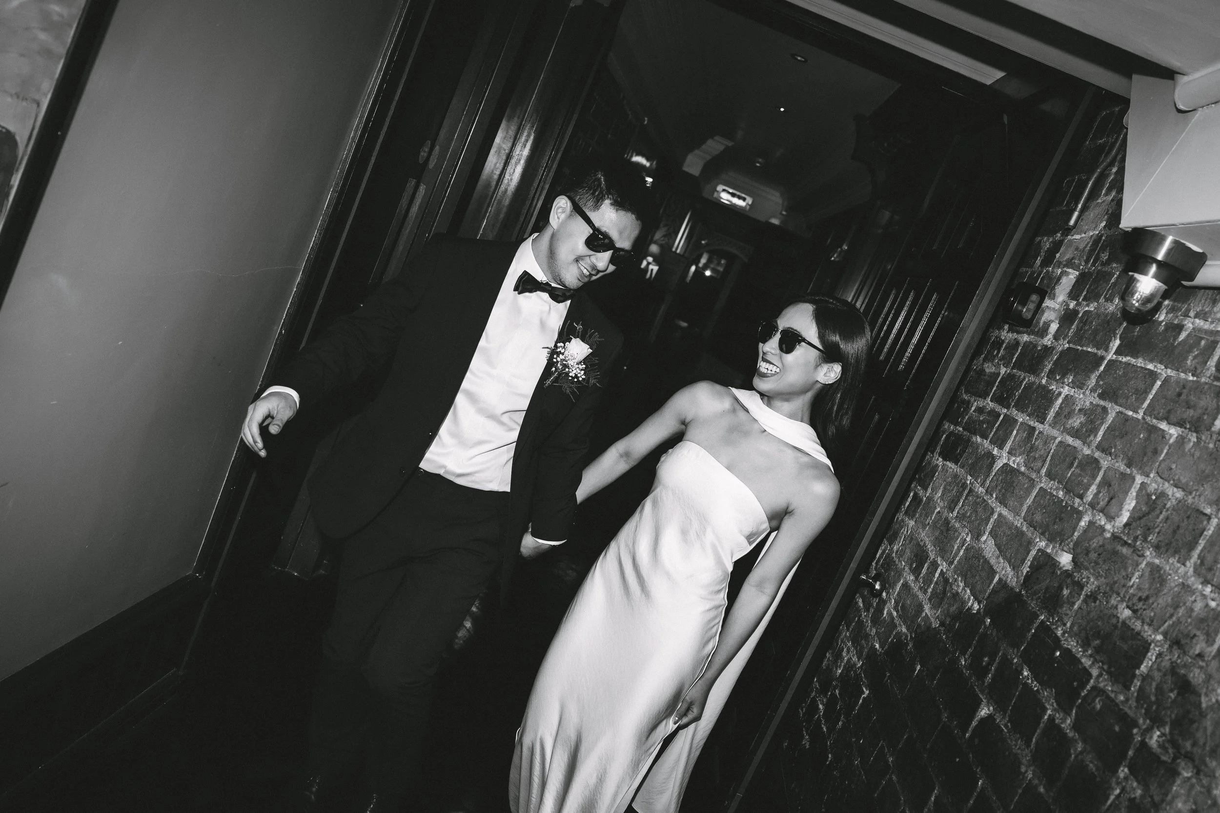 A man and woman in formal attire and sunglasses are smiling and holding hands, walking through a corridor with brick walls in black and white.