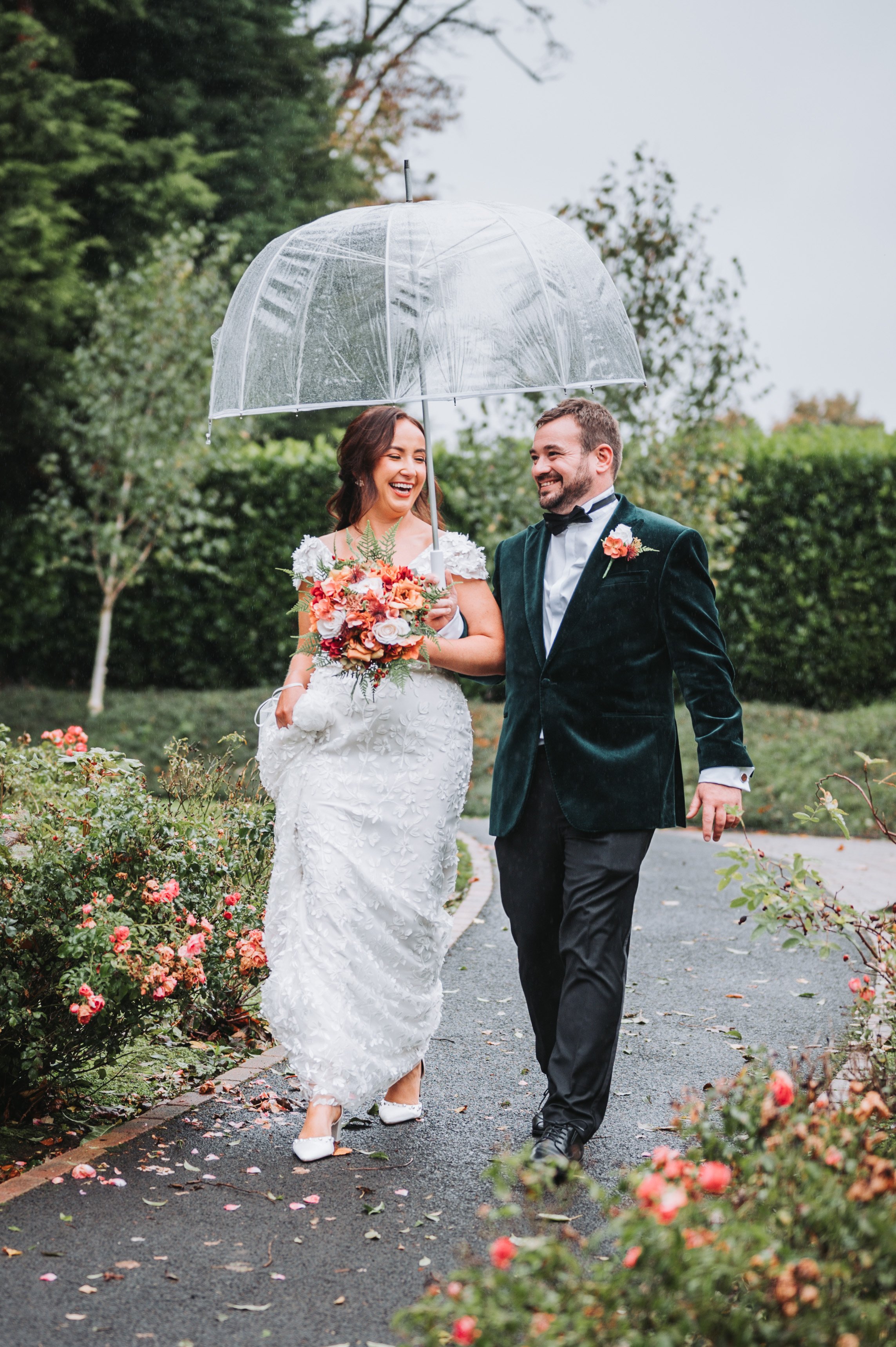 A smiling bride and groom walk outside on a rainy day under a transparent umbrella, surrounded by pink and red roses, with greenery in the background.