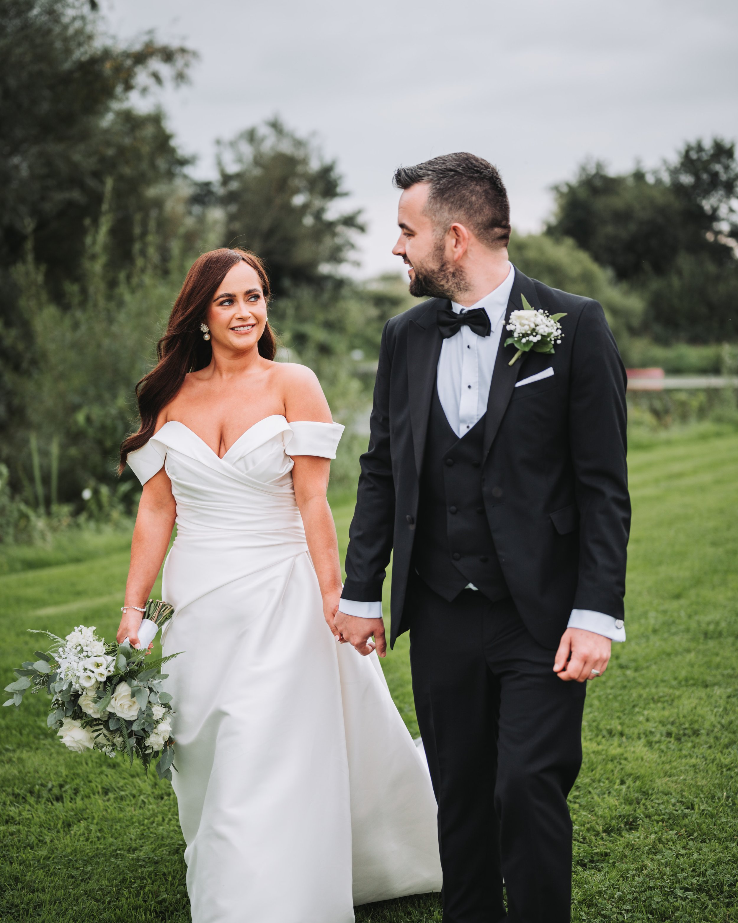 Bride and groom walking hand in hand outdoors, smiling at each other. She is wearing an off-shoulder white wedding gown and holding a bouquet, he is wearing a black tuxedo with a bow tie and boutonniere.