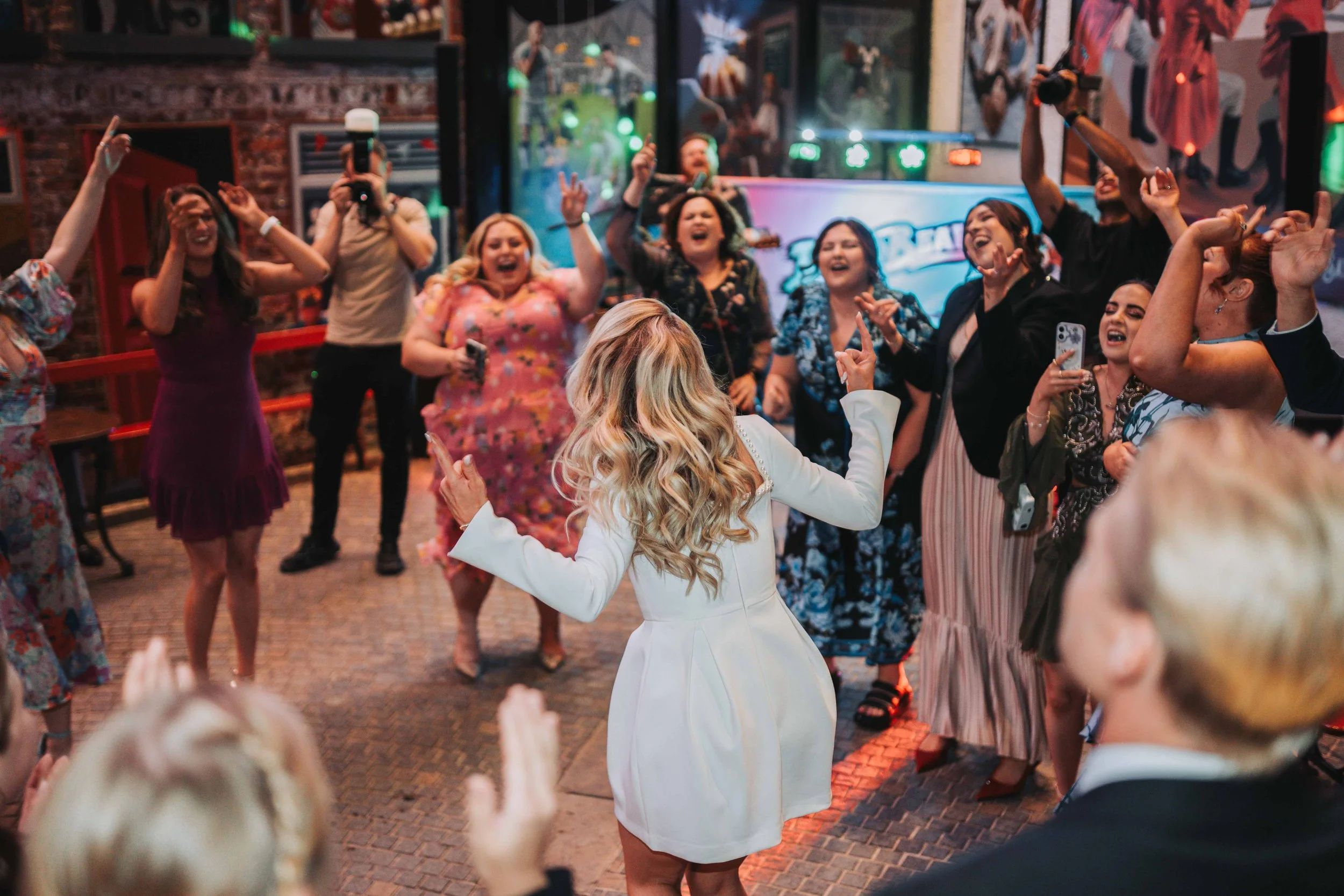 A group of people dancing and celebrating at a party or celebration, with a woman in a white dress dancing in the center, surrounded by friends taking photos and cheering.