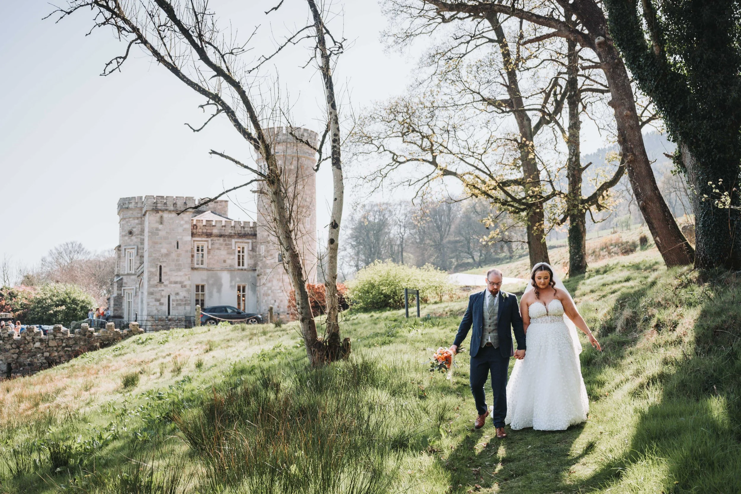 A bride and groom walking hand-in-hand on a grassy path in a park, with a historic stone castle building in the background, surrounded by leafless trees and a bright, sunny sky.