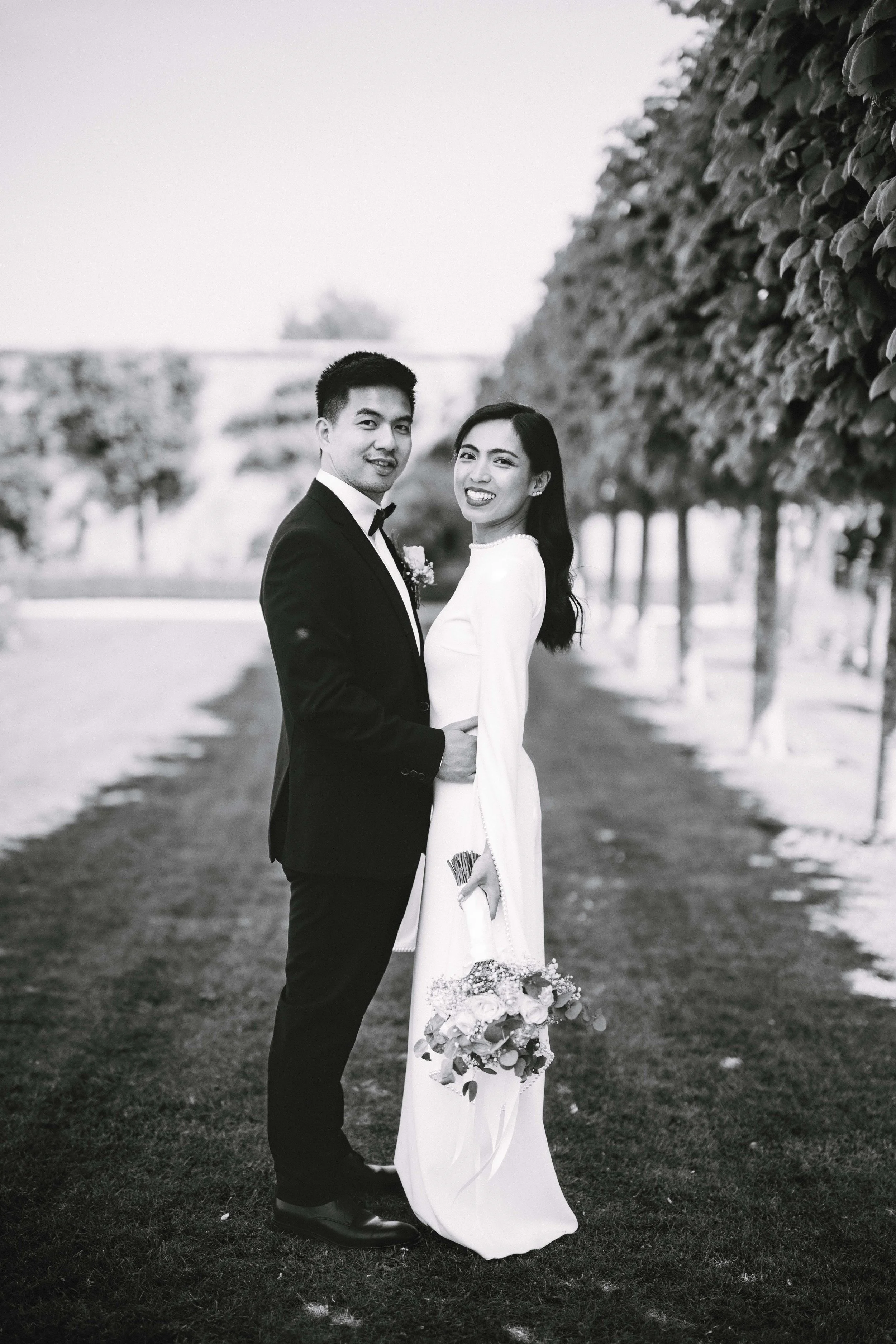 Black and white photo of a bride and groom standing on a grassy path lined with trees, smiling at the camera, the bride holding a bouquet of flowers.