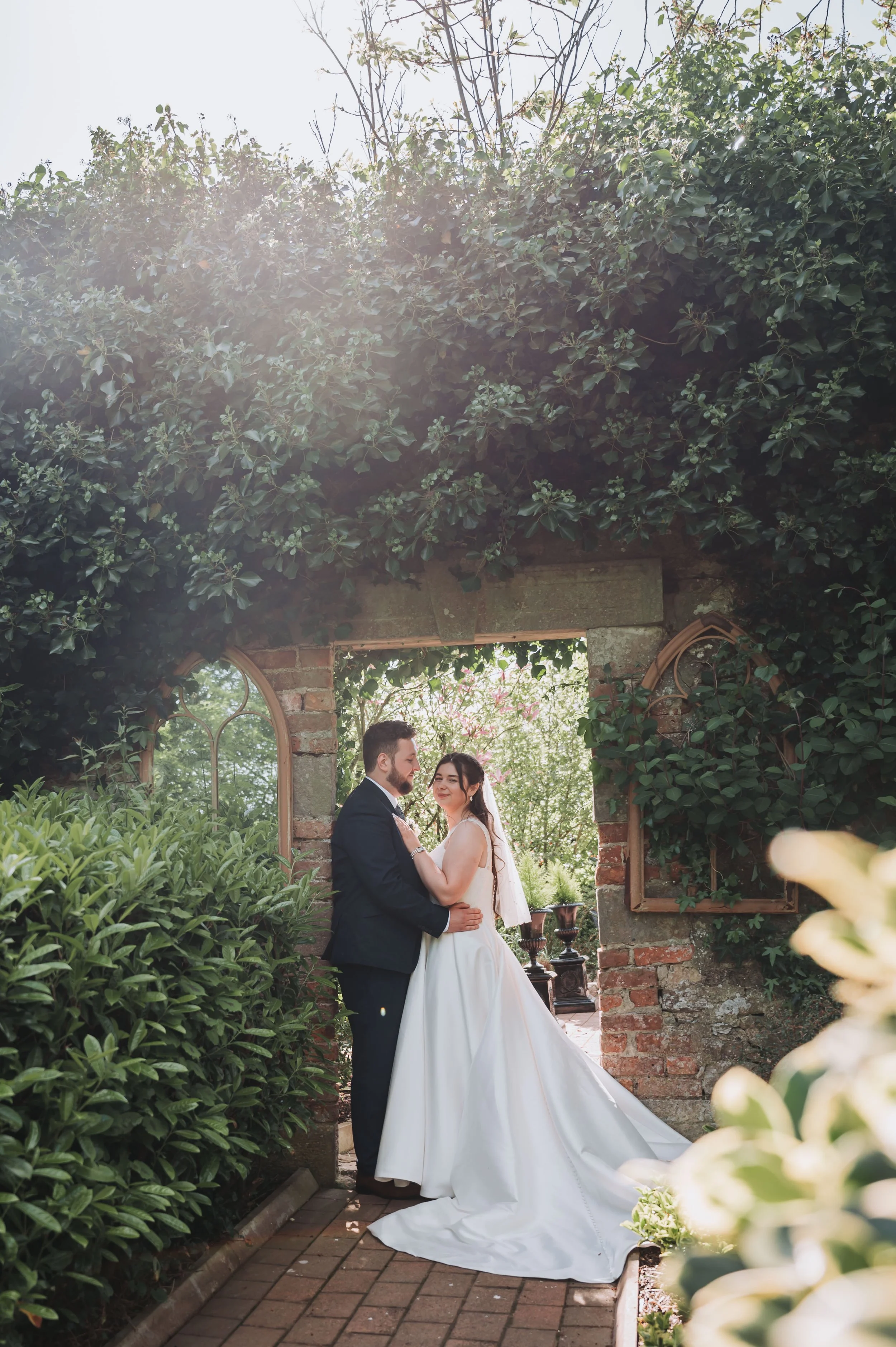 A bride and groom standing in a garden archway, embracing each other on their wedding day, surrounded by lush greenery and blooming flowers.
