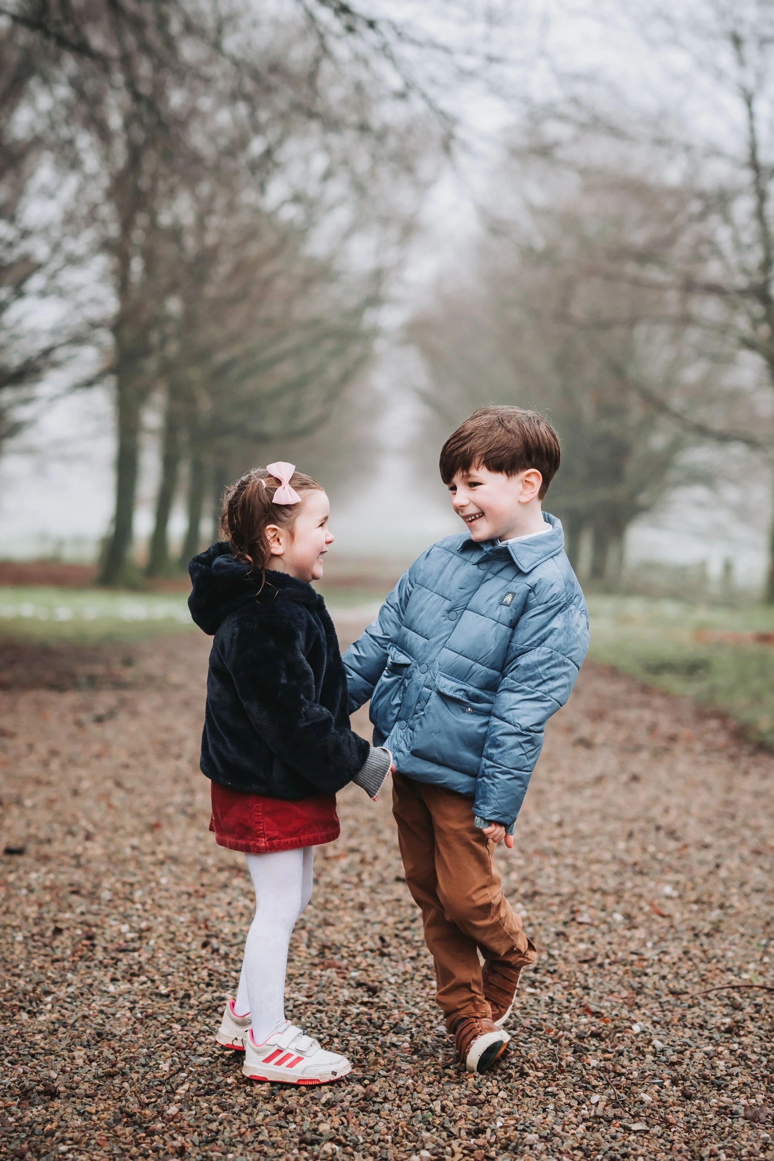 A young boy and girl are holding hands and smiling at each other while standing on a gravel path outdoors, with trees and an overcast sky in the background.