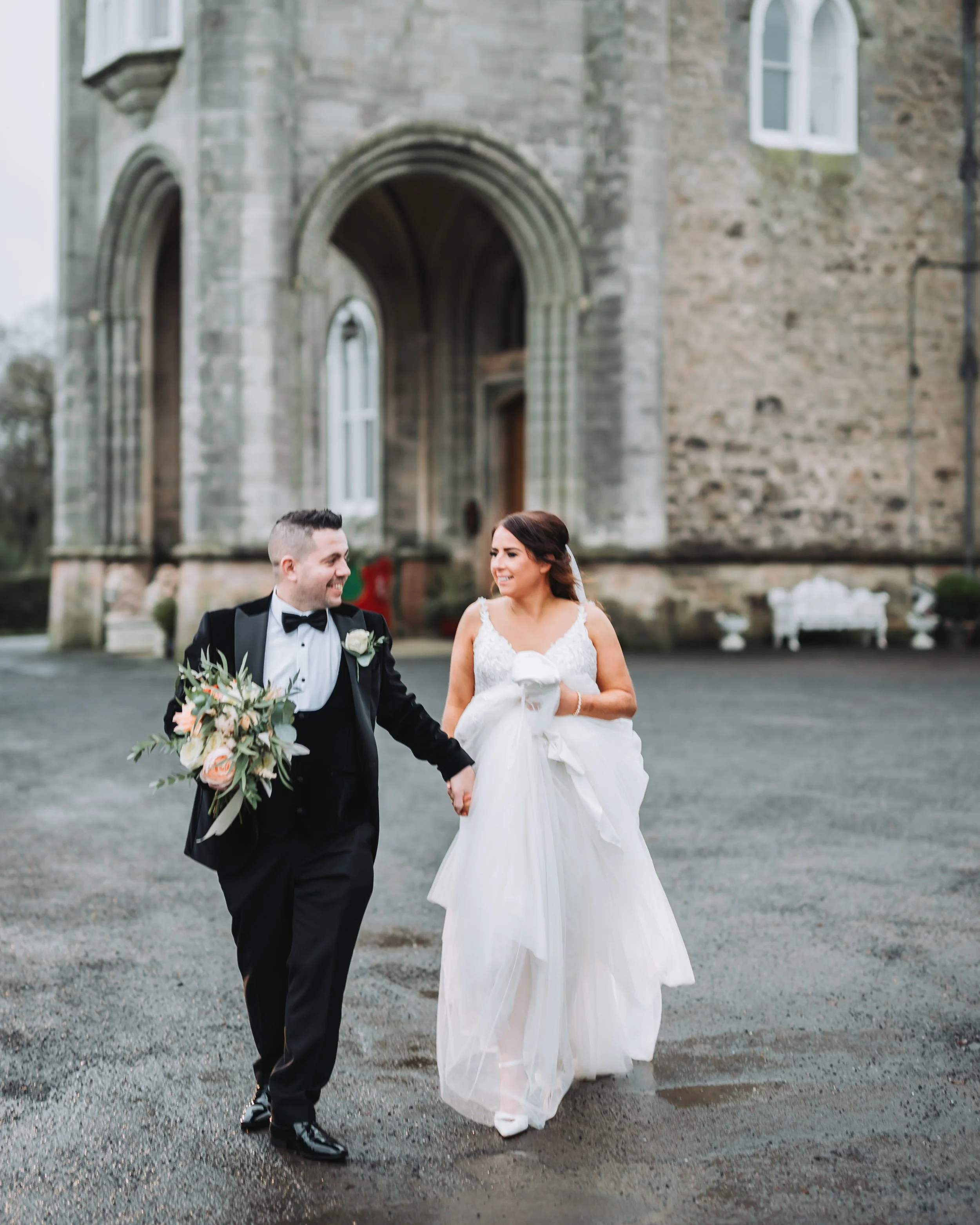 A bride and groom walking hand-in-hand outside a stone building, smiling at each other. The groom is wearing a black tuxedo with a bow tie and holding a bouquet of flowers. The bride is in a white wedding dress, holding her skirt as they walk.