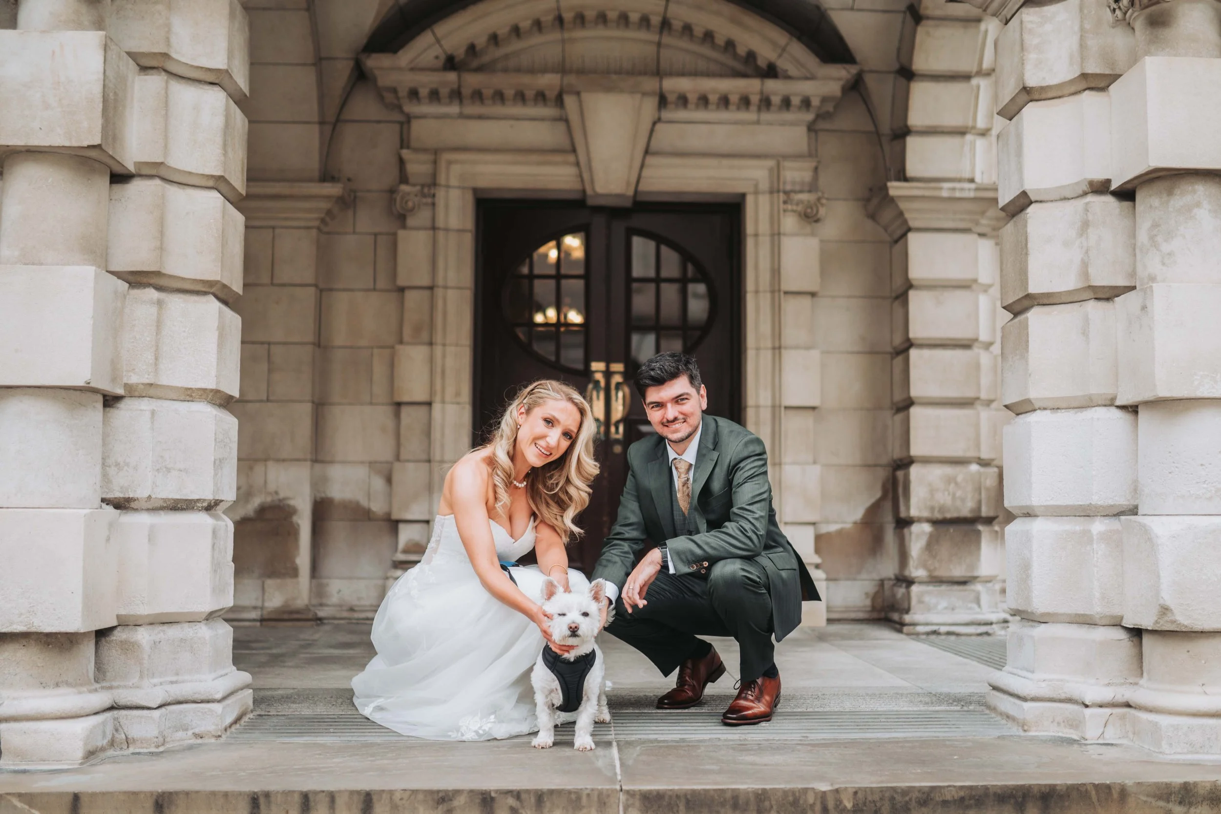 A bride and groom in wedding attire crouch with a small white dog on a historic stone building steps, smiling at the camera.