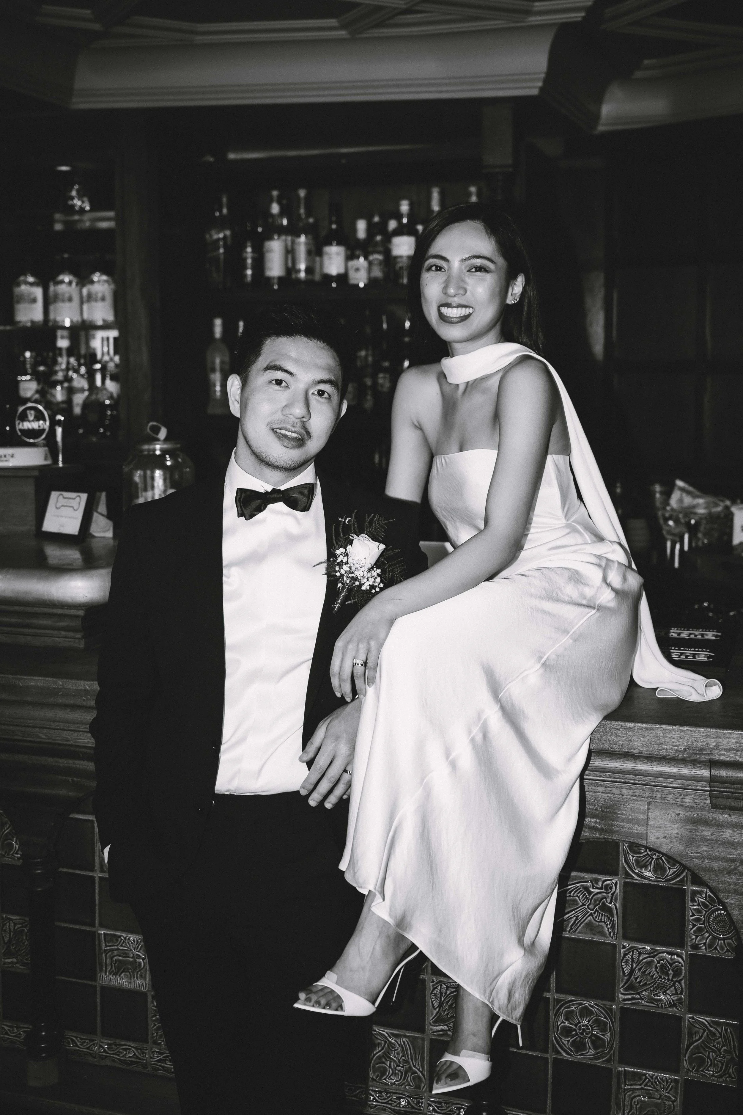 A black-and-white photo of a man in a tuxedo with a bow tie and a boutonniere, and a woman in a strapless dress and high heels, sitting at a bar with liquor bottles in the background.