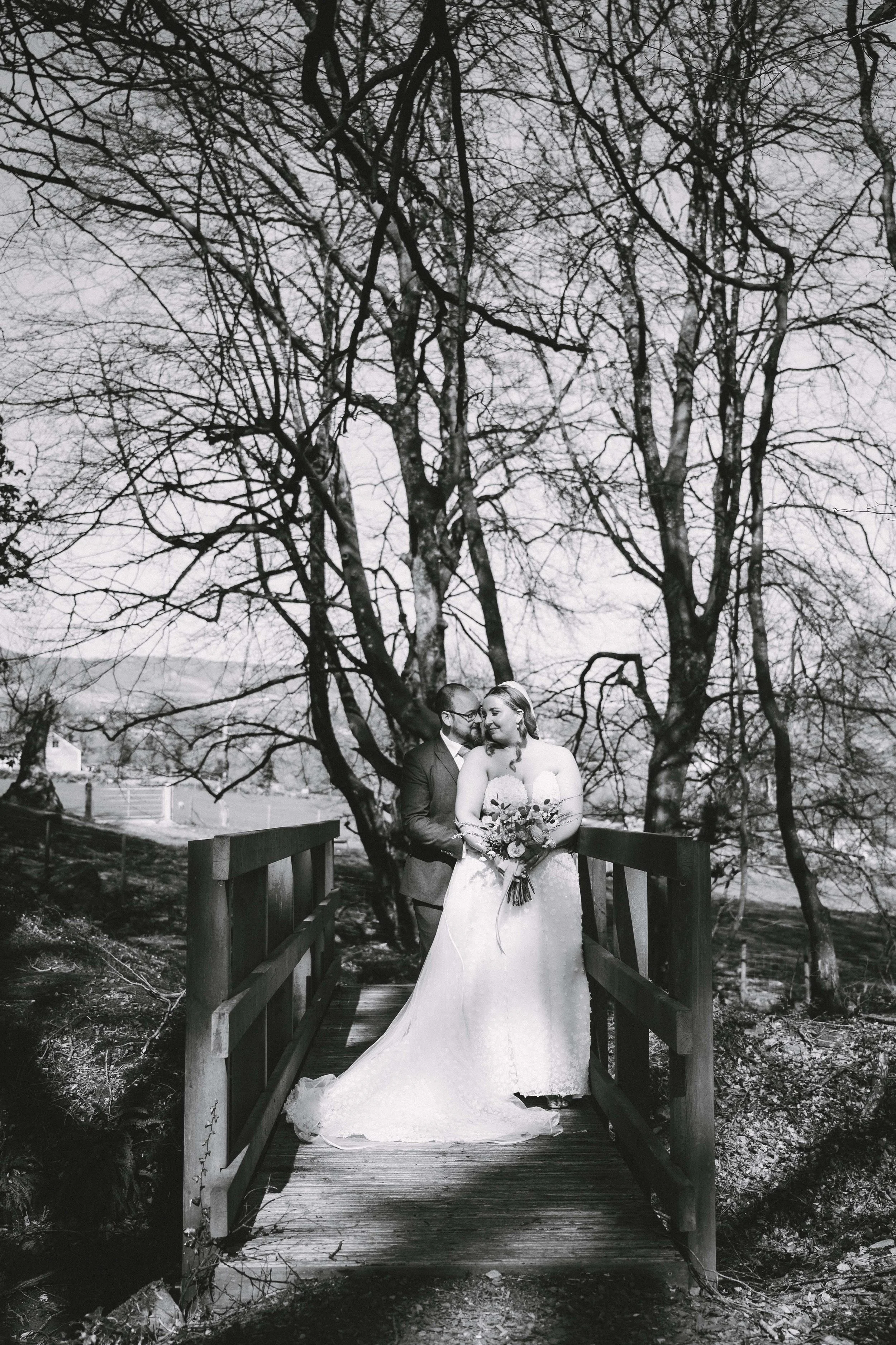 Black and white photograph of a bride and groom standing on a wooden bridge outdoors, with leafless trees and open land in the background. The bride wears a strapless wedding gown and holds a bouquet, while the groom is in a suit, and they are leanin