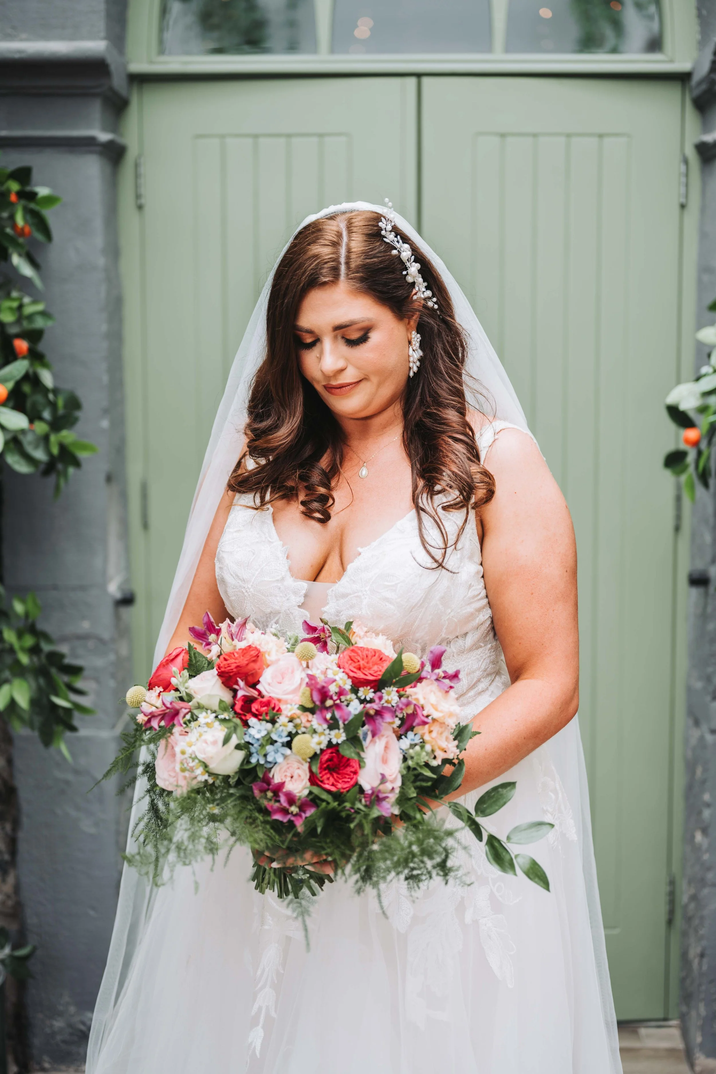 A bride with brown curly hair, wearing a white wedding gown and veil, holding a colorful bouquet of pink, red, purple, and white flowers, standing in front of a green door surrounded by greenery.