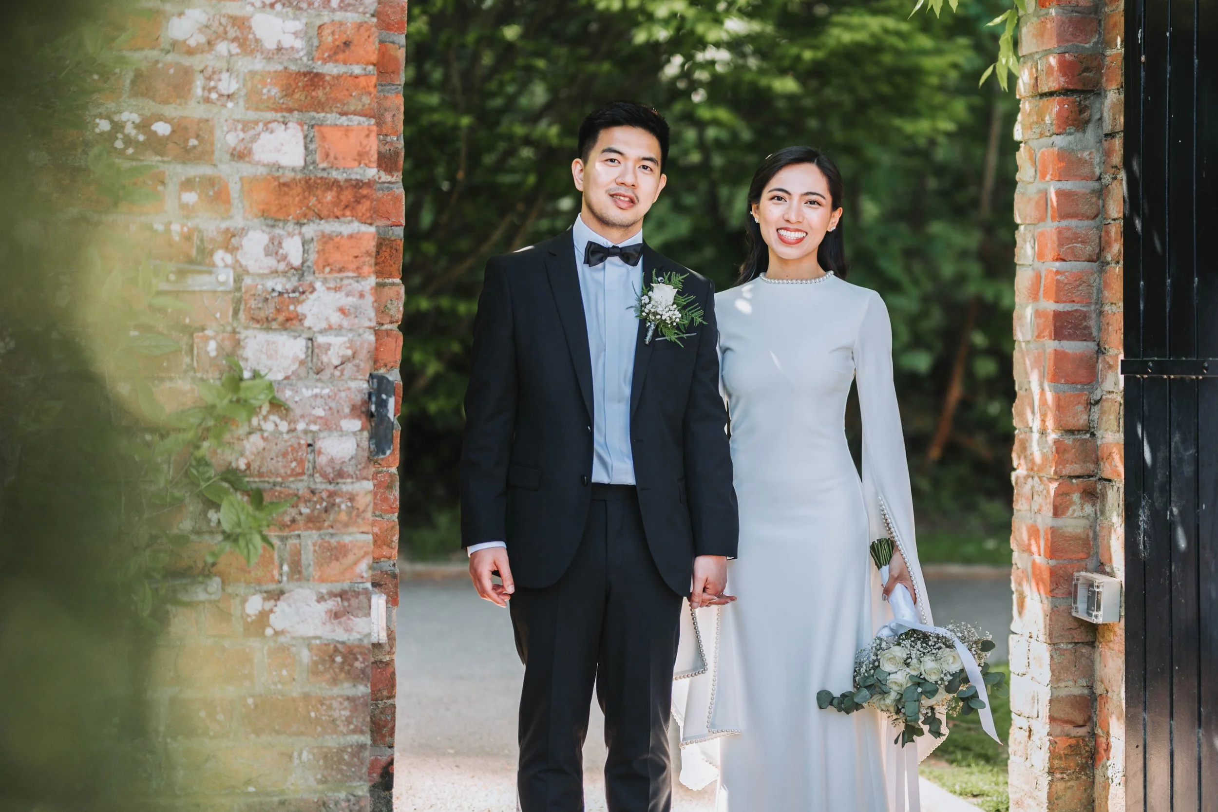 A bride and groom standing outdoors near a brick wall, smiling at the camera. The groom is dressed in a black suit with a bow tie and boutonnière, holding a glass. The bride is wearing a long white wedding dress with pearl details, holding a bouquet 