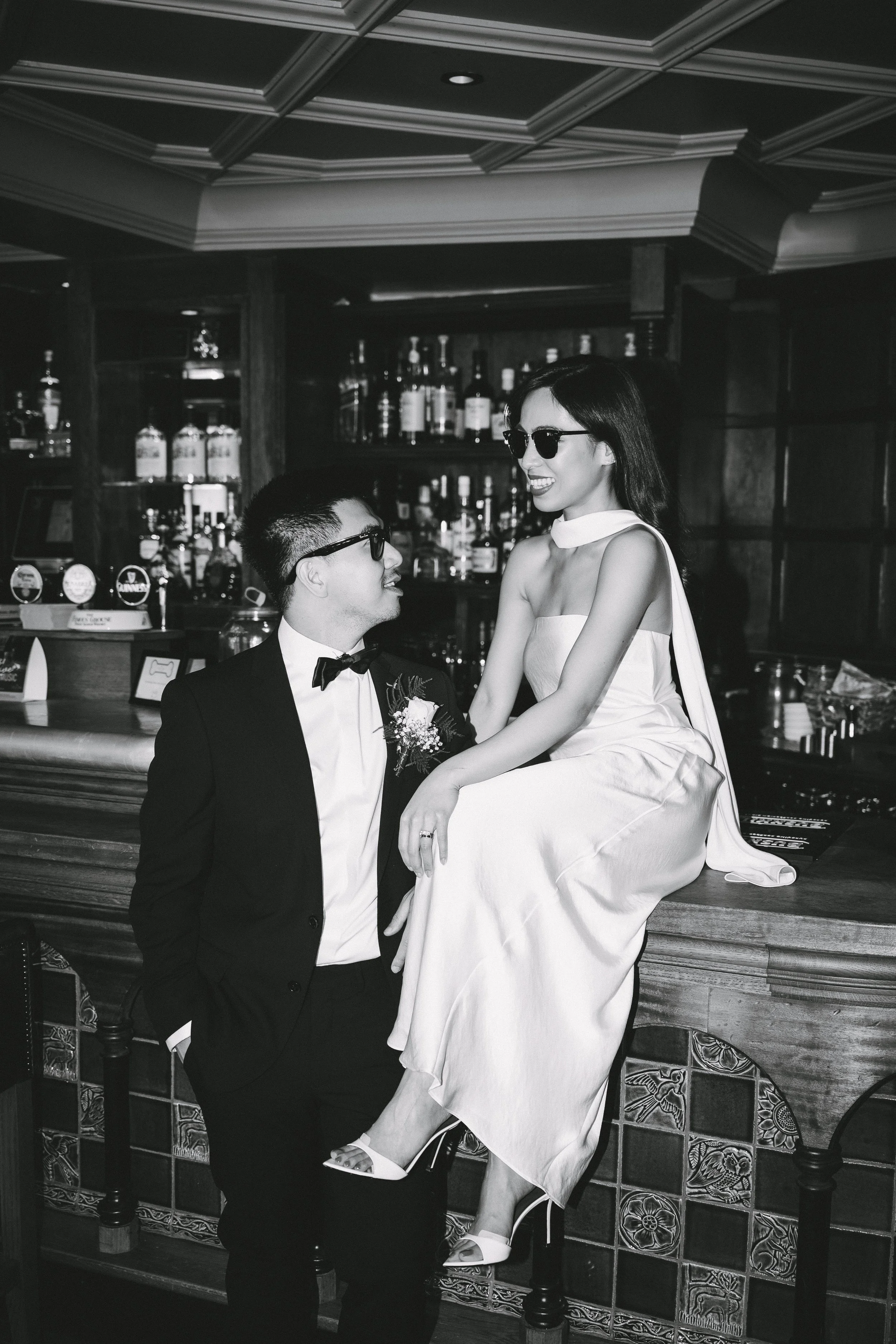 A black and white photo of a couple at a bar, dressed in formal attire. The woman is sitting on the bar counter wearing a white dress and high heels, and the man is standing next to her in a tuxedo with a bow tie, looking up at her. They are both smi