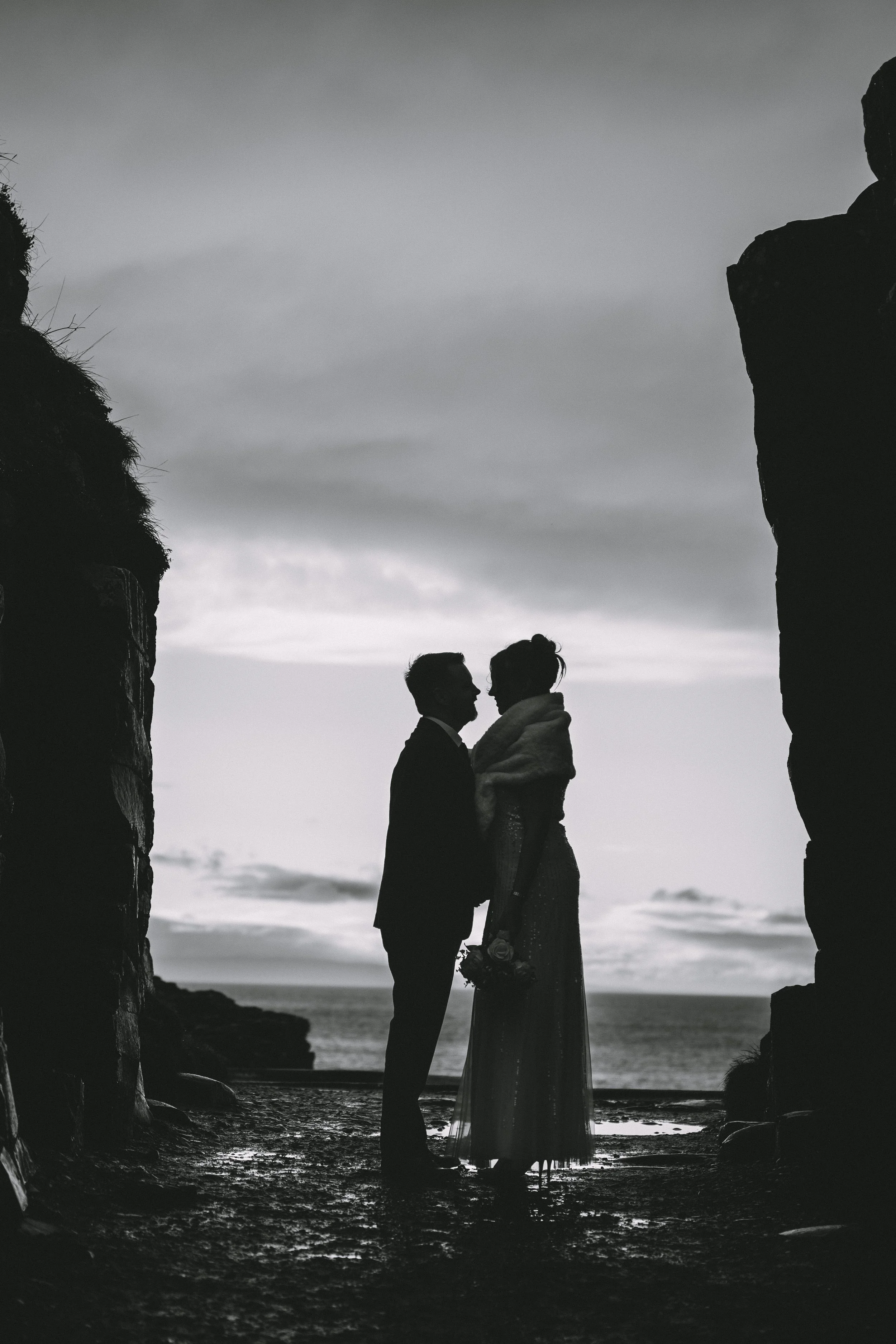 Silhouette of a couple, possibly newlyweds, standing close together at a beachside cliff during sunset, with the ocean in the background.