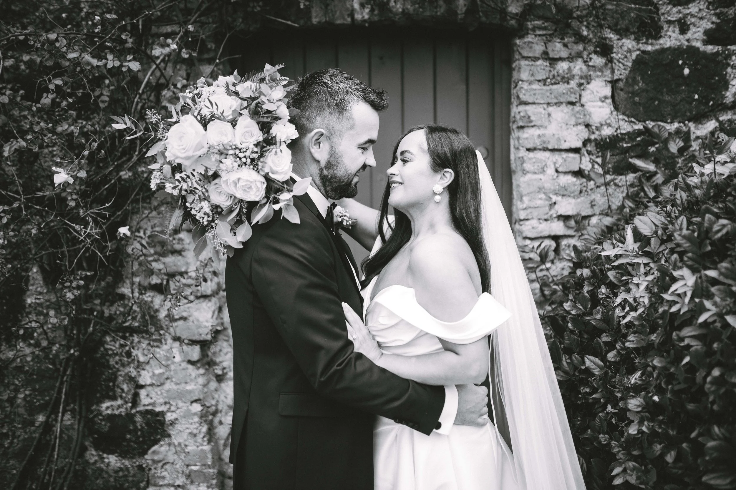 A black-and-white photo of a bride and groom in wedding attire, smiling and looking into each other's eyes, with the groom holding a bouquet of flowers and standing against a rustic brick wall with foliage.