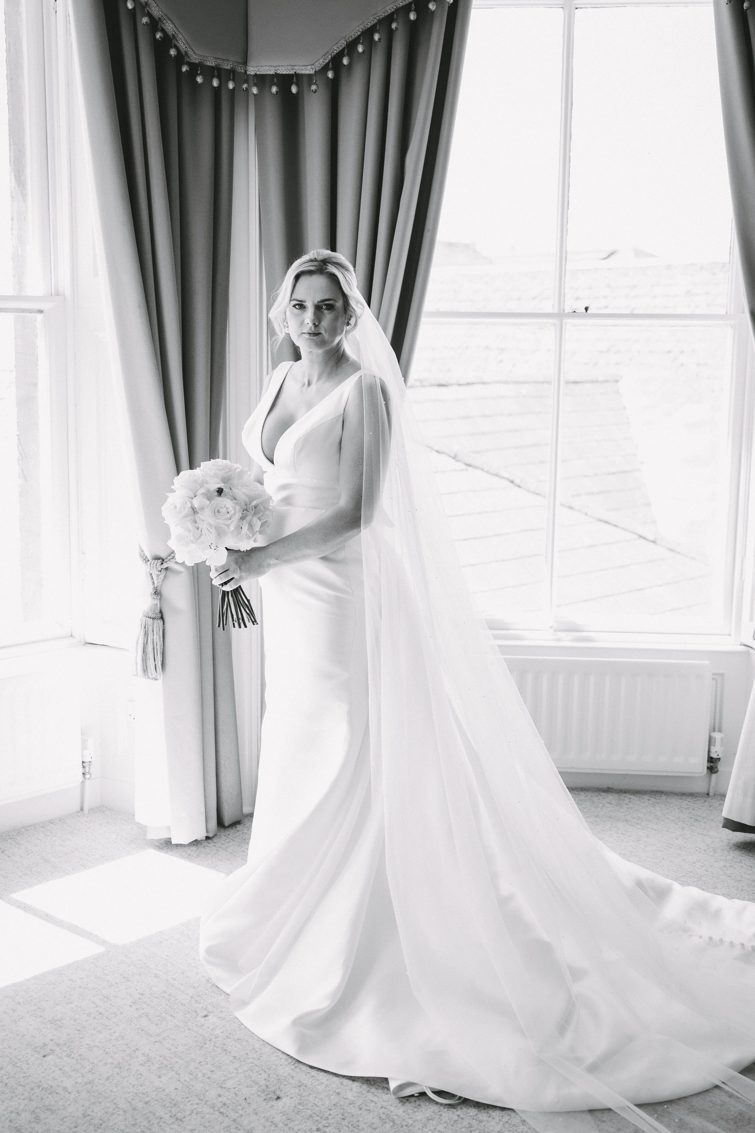 Black and white photo of a bride in a wedding dress holding a bouquet of roses, standing near a window with drapes.