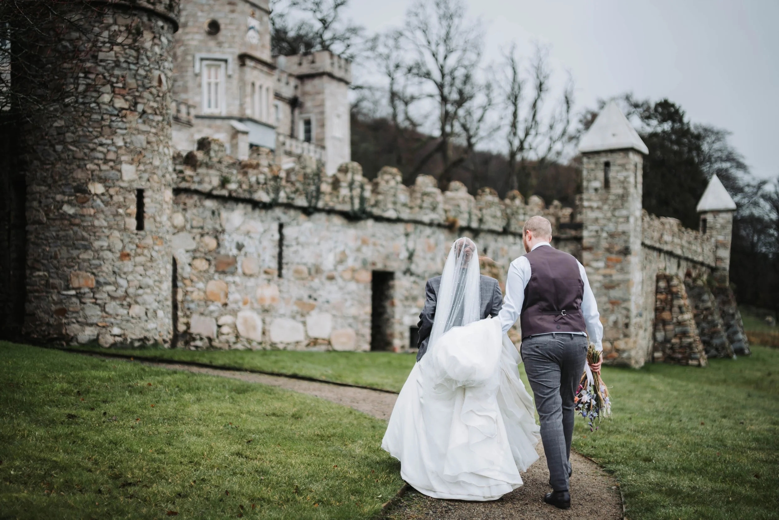 Bride and groom walking towards a stone castle, bride in white wedding dress and veil, groom in grey suit vest and trousers, holding a bouquet of flowers, during daytime.