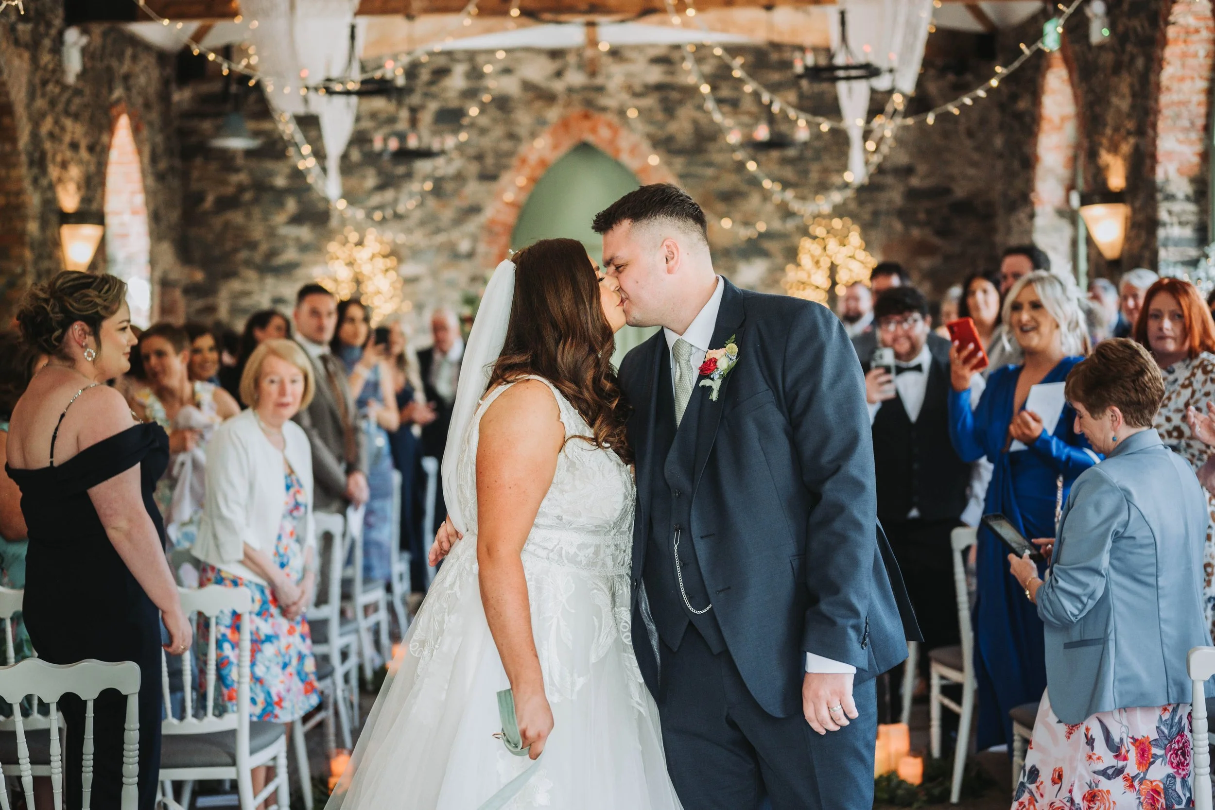 A bride and groom kiss at their wedding ceremony in a rustic venue with stone walls and string lights, surrounded by seated and standing guests.