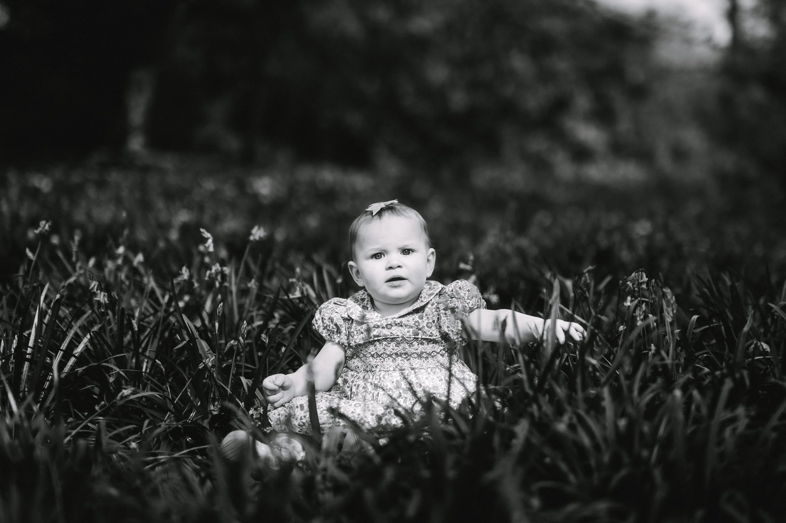 Black and white photo of a young girl sitting in tall grass outdoors, looking confused or curious.