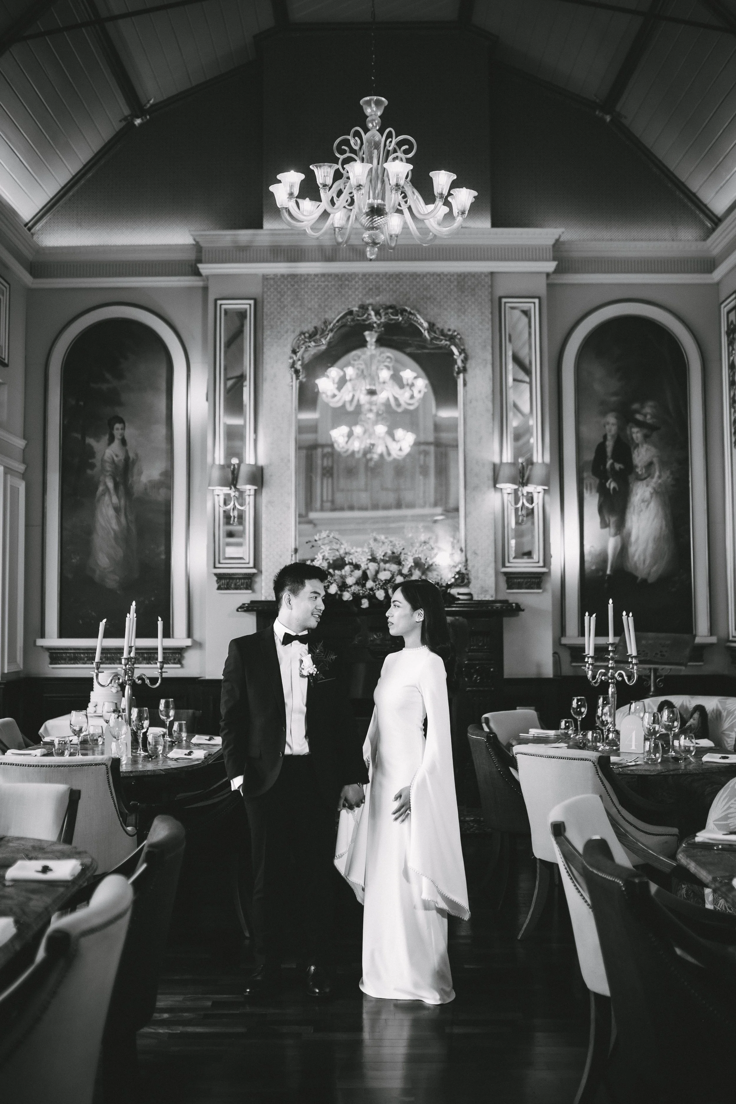 Black and white photo of a bride and groom standing hand in hand in an elegant, decorated wedding reception hall with chandeliers and paintings.